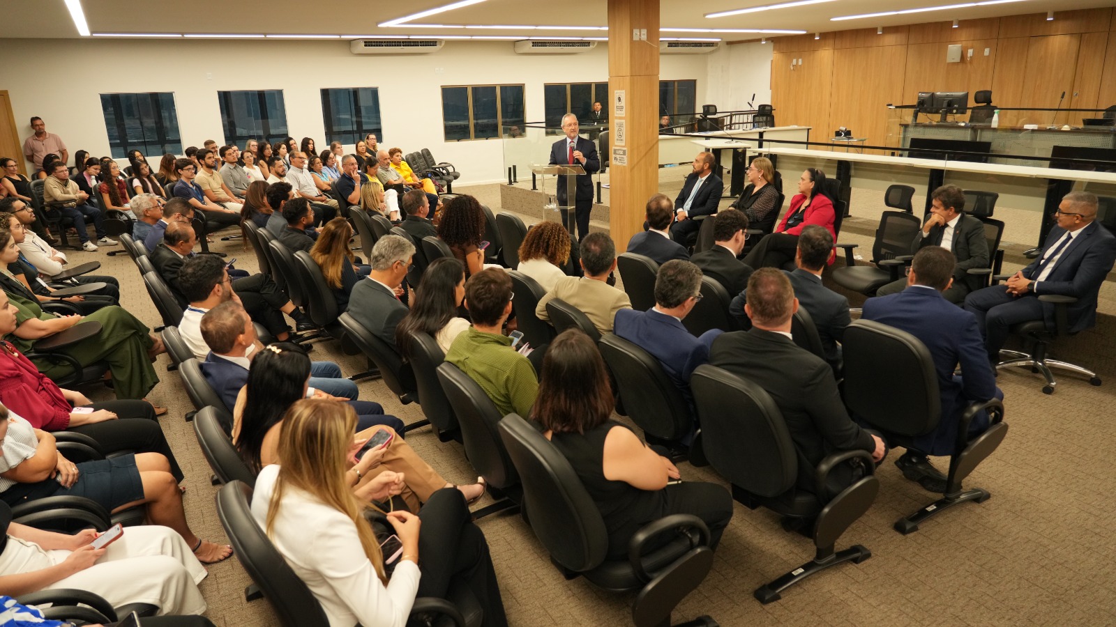 A imagem mostra uma sala do Tribunal do Júri, com várias pessoas sentadas em cadeiras organizadas em fileiras, assistindo à abertura oficial das Correições Gerais Ordinárias, em Palmas. À frente, o corregedor-geral da Justiça, Pedro Nelson de Miranda Coutinho, de terno, discursa em um púlpito, enquanto autoridades e convidados acompanham a solenidade.