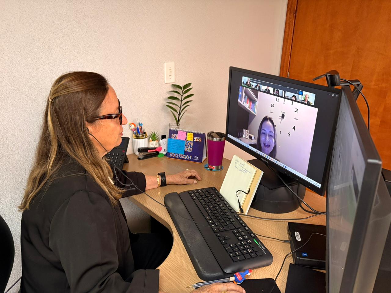 Desembargadora Maysa em gabinete participa de videoconferência pelo computador, com teclado e materiais de escritório sobre a mesa. Na tela, aparecem os participantes da videoconferência. 