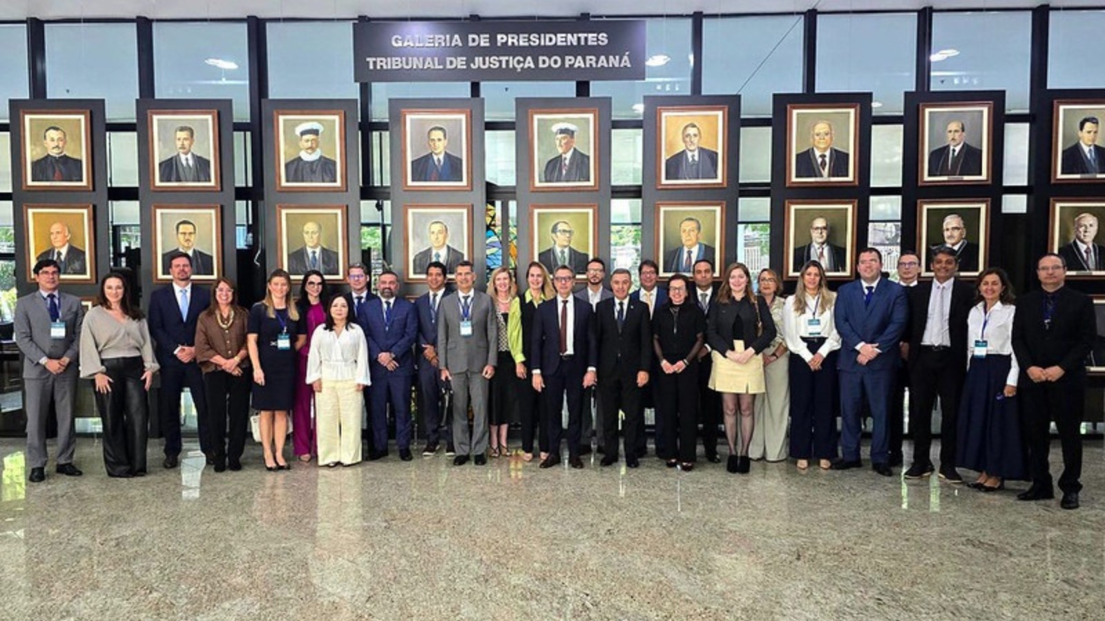 A imagem mostra um grupo de autoridades, magistrados(as) e servidores(as) posa para foto institucional no hall do Tribunal de Justiça do Paraná. Ao fundo, está a Galeria de Presidentes do Tribunal de Justiça do Paraná, com retratos emoldurados dos antigos dirigentes da instituição expostos em uma parede de vidro. Os participantes estão alinhados lado a lado, em traje formal, em frente à galeria.