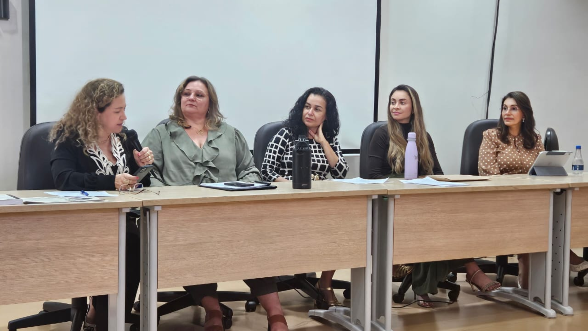 A imagem mostra cinco mulheres sentadas lado a lado em uma mesa de debate, durante o evento Colóquio Internacional Psicanálise e Crítica Social do Trabalho. Uma das participantes fala ao microfone enquanto as demais acompanham atentamente a discussão.
