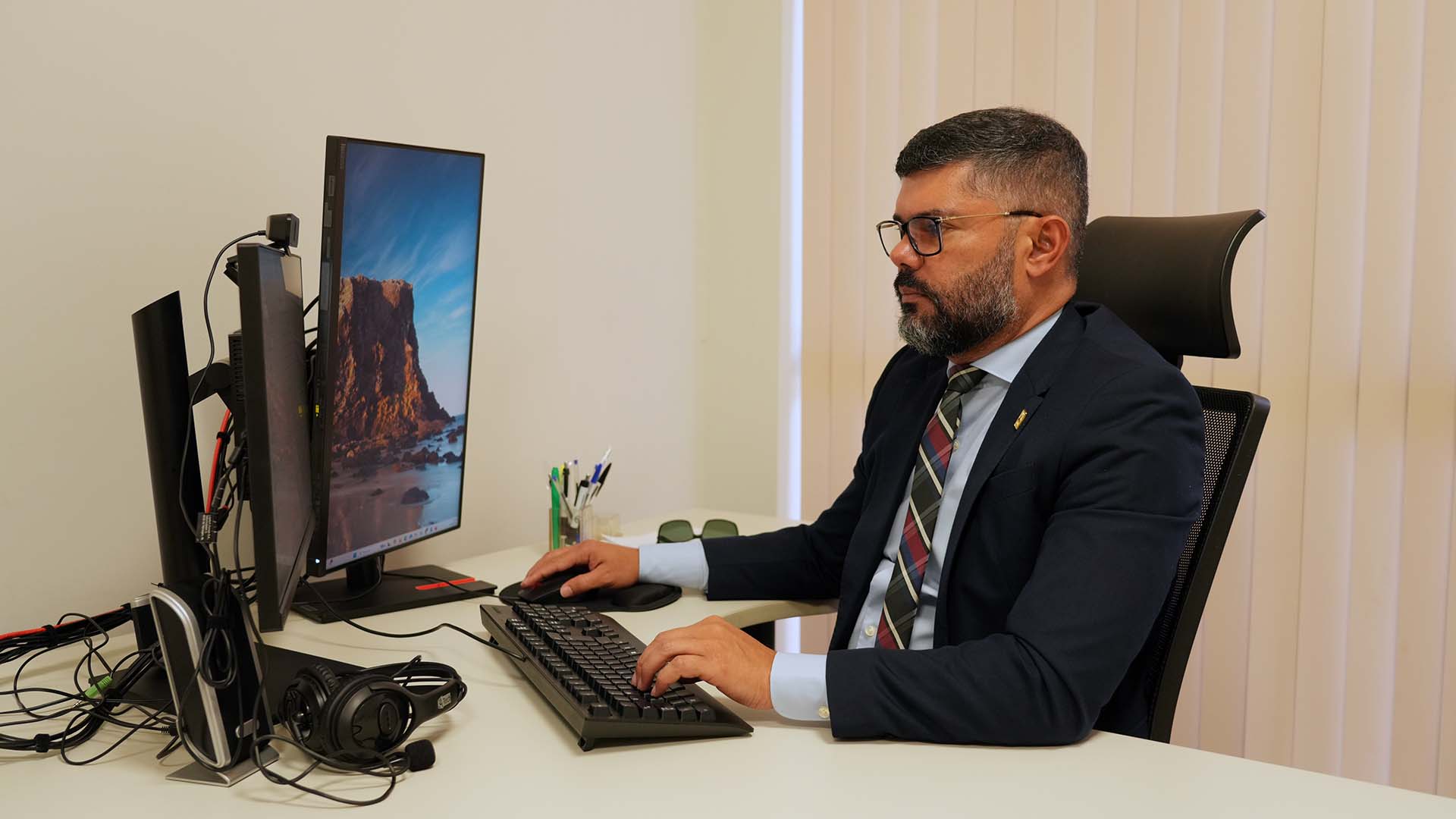 O juiz Wellington Magalhães aparece sentado em sua estação de trabalho, em ambiente de gabinete, concentrado diante de dois monitores de computador. Vestindo terno escuro, camisa clara e gravata, o magistrado utiliza teclado e mouse enquanto analisa informações na tela. Sobre a mesa, há materiais de escritório, como canetas, óculos e um headset, além de cabos e equipamentos do computador. Ao fundo, persianas verticais claras compõem o ambiente institucional.