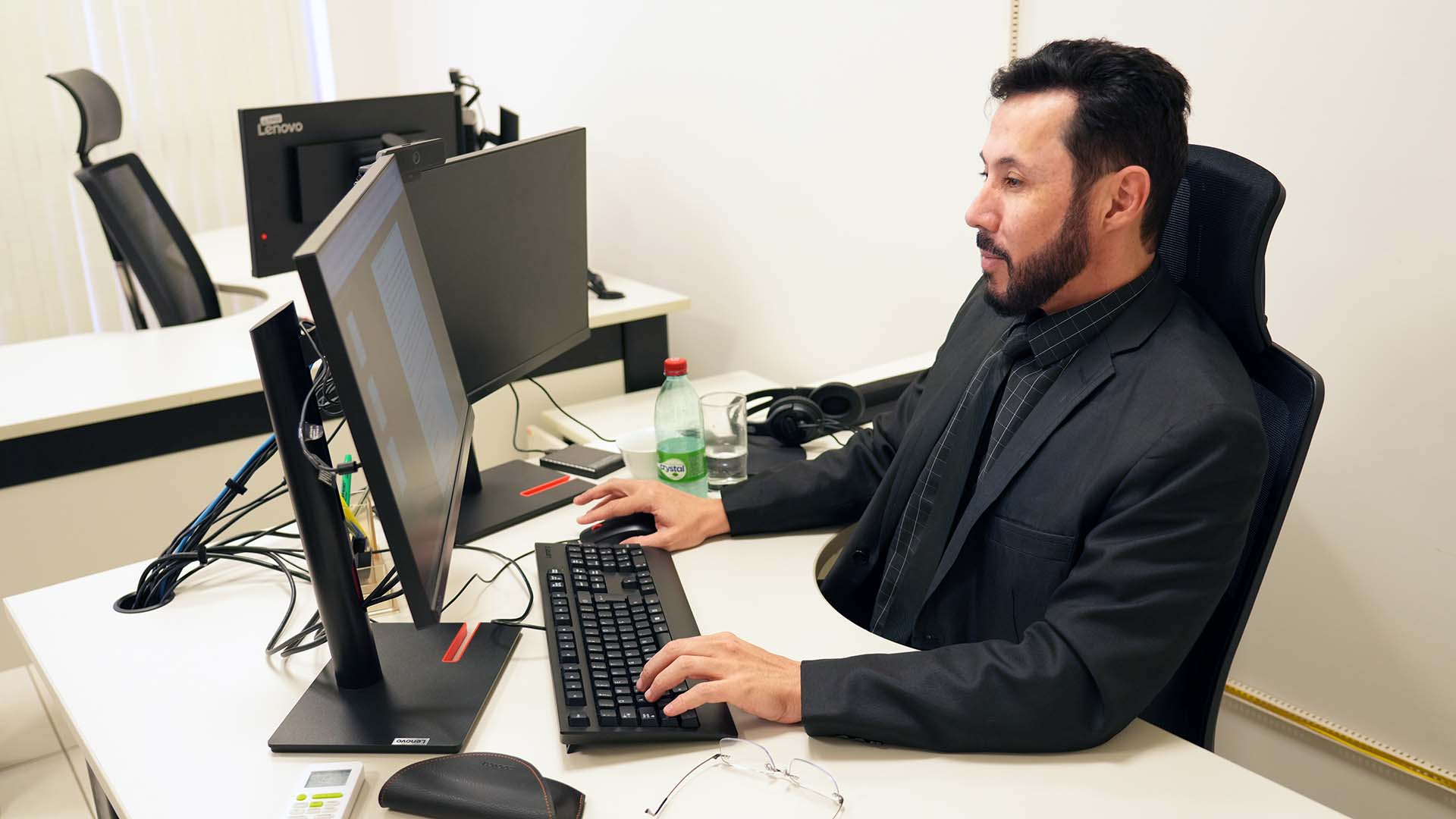 O juiz Fabiano Gonçalves aparece sentado em sua estação de trabalho, em ambiente de gabinete, diante de dois monitores de computador. Vestindo terno escuro e camisa preta, o magistrado utiliza teclado e mouse enquanto analisa informações na tela. Sobre a mesa estão objetos de uso cotidiano, como um par de óculos, fones de ouvido, garrafa de água e copo. Ao fundo, outras estações de trabalho com computadores compõem o espaço institucional.