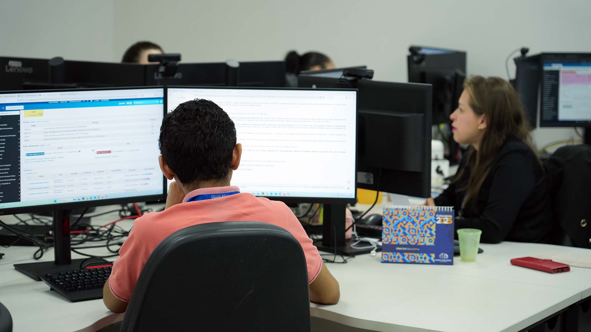 Ambiente de trabalho institucional com servidores do Nacom atuando em computadores de mesa. Em primeiro plano, um servidor, visto de costas, utiliza dois monitores enquanto analisa informações no sistema. Ao lado, uma servidora também trabalha diante do computador. Ao fundo, outros profissionais aparecem em suas estações. Na mesa, há objetos de uso cotidiano, como teclado, mouse, copo plástico e um calendário de mesa.