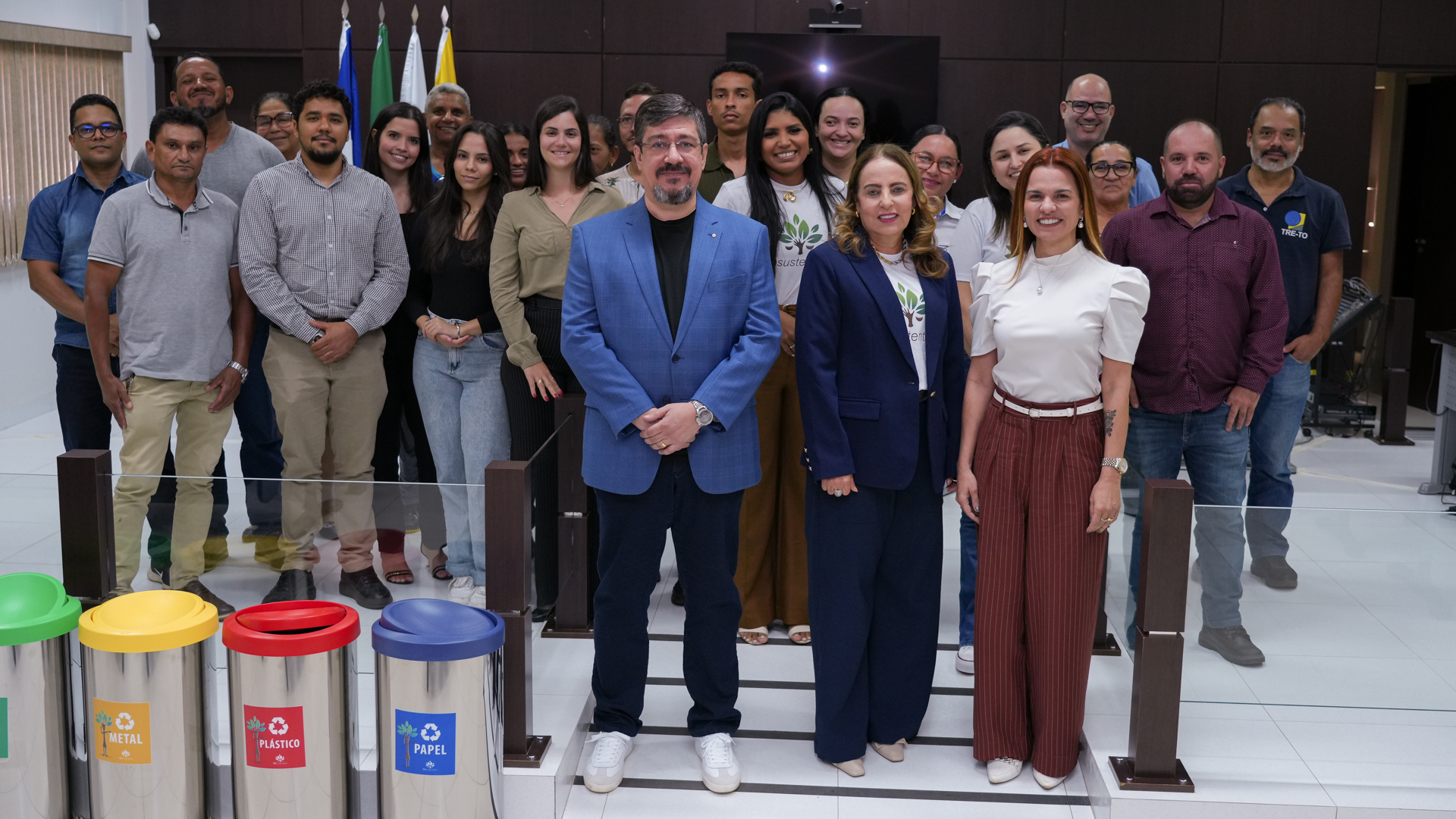 A imagem mostra um grupo de cerca de 20 pessoas, principalmente adultos, posando juntas em um ambiente interno, provavelmente um escritório. No centro, há duas pessoas em trajes formais: um homem de terno azul e uma mulher com blazer escuro. Eles estão rodeados por outras pessoas que também parecem profissionais e estão sorrindo. À frente, há três lixeiras de reciclagem, identificadas como "METAL", "PLASTICO" e "PAPEL", sugerindo um foco em sustentabilidade. O ambiente é bem iluminado e organizado, transmitindo uma sensação de colaboração e responsabilidade ambiental.