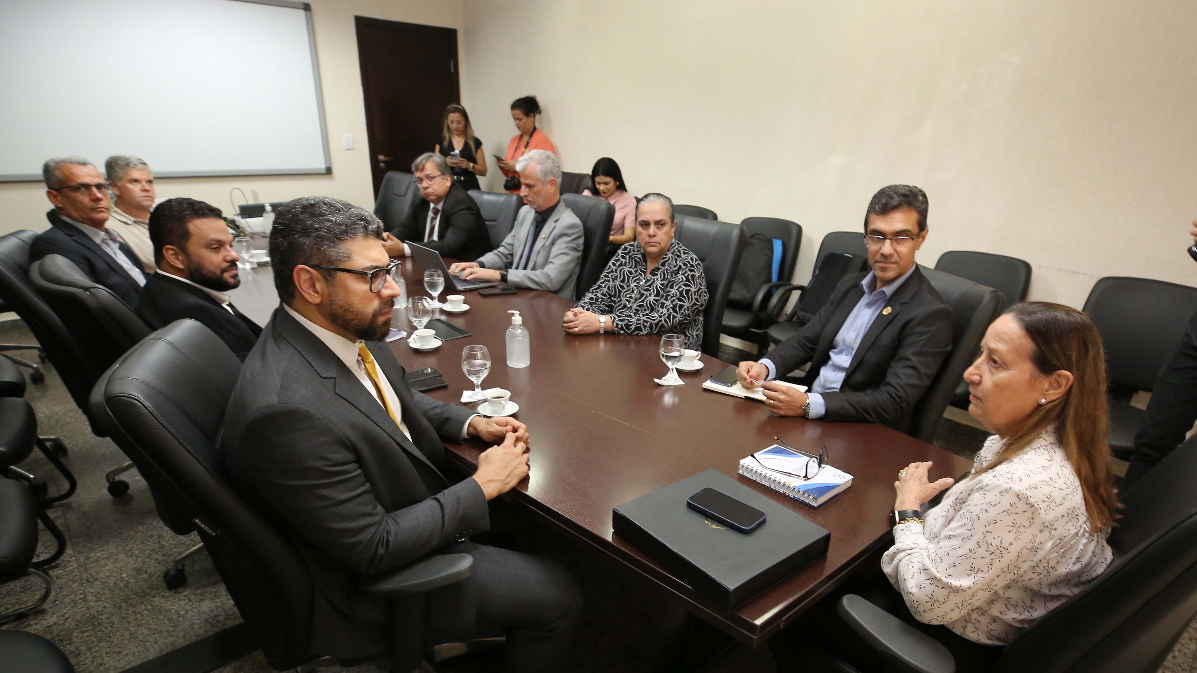 A imagem mostra um grupo de pessoas sentadas em torno de uma mesa grande e retangular, em uma sala. Há pelo menos 10 pessoas à mesa.  As pessoas estão vestidas com roupas formais.