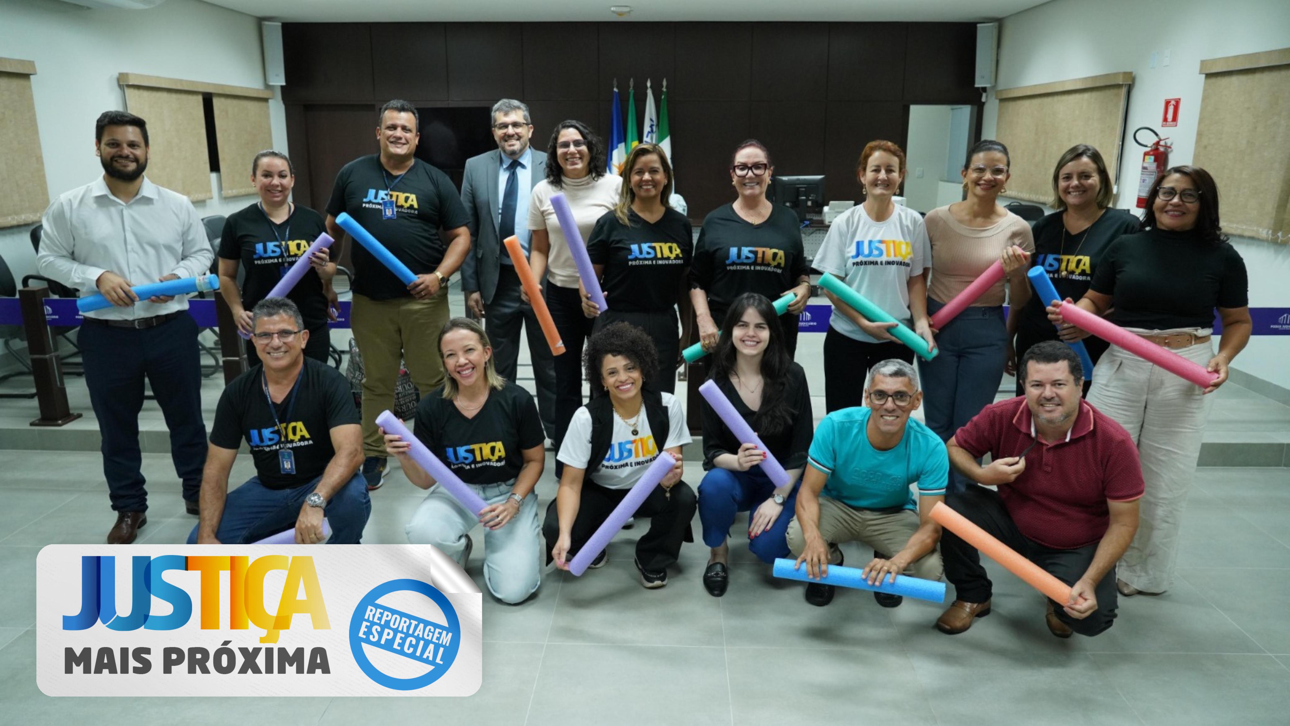 A imagem mostra um grupo de 17 pessoas posando para uma foto em um ambiente interno iluminado. Algumas pessoas estão de pé, outras agachadas, e todas sorriem para a câmera. Elas seguram rolos coloridos de espuma, usados em atividades. Algumas vestem camisetas pretas ou brancas com a palavra "Justiça" em destaque. Ao fundo, há mesas, cadeiras e bandeiras.