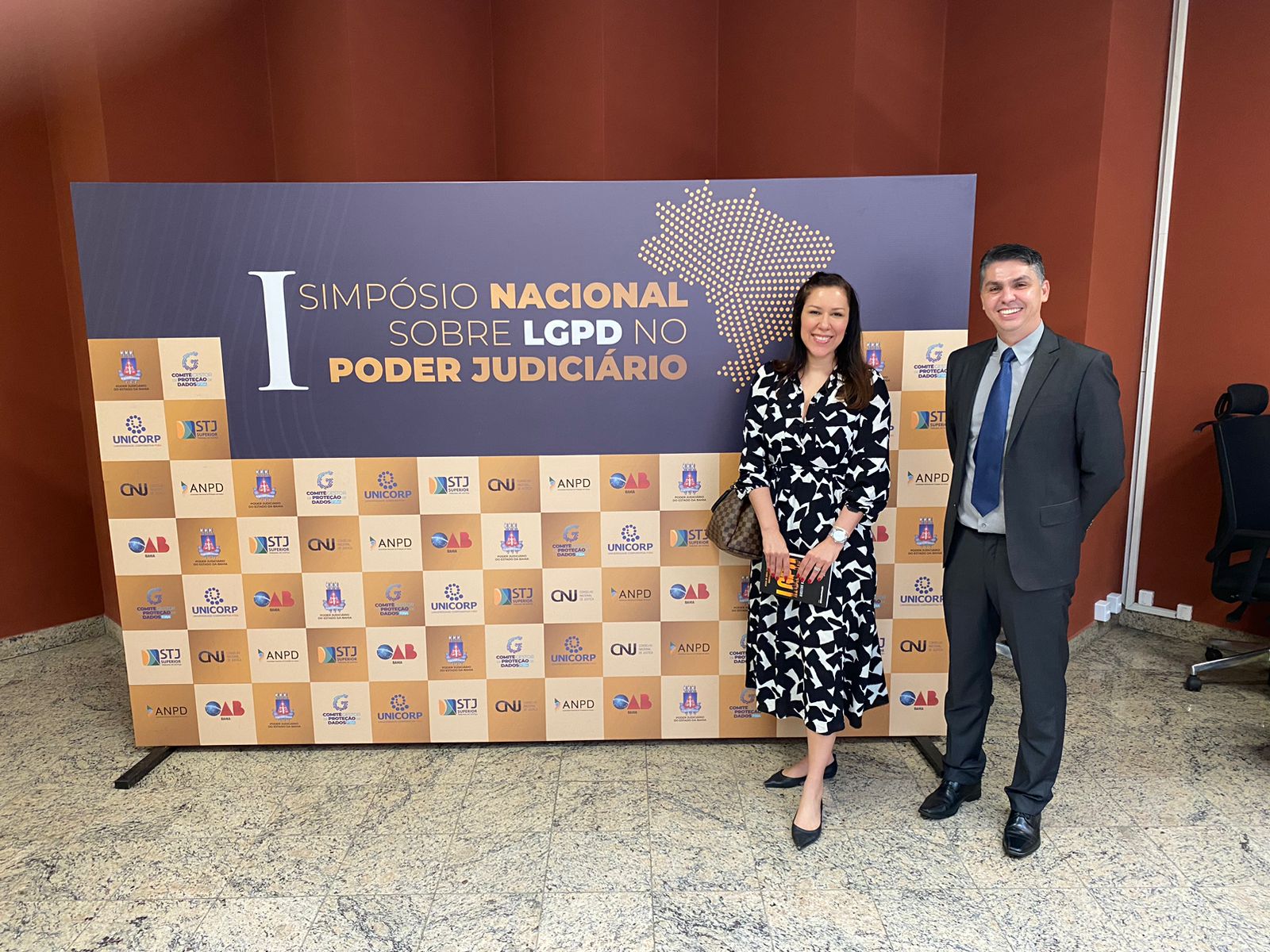 Delegada Milena, uma mulher de cabelos longos e pretos usando um vestido estampado em preto e branco, em pé ao lado de Alex, vestido de terno cinza, camisa azul claro e grata azul royal, também em pé à frente de uma placa com o nome do evento e logos dos organizadores