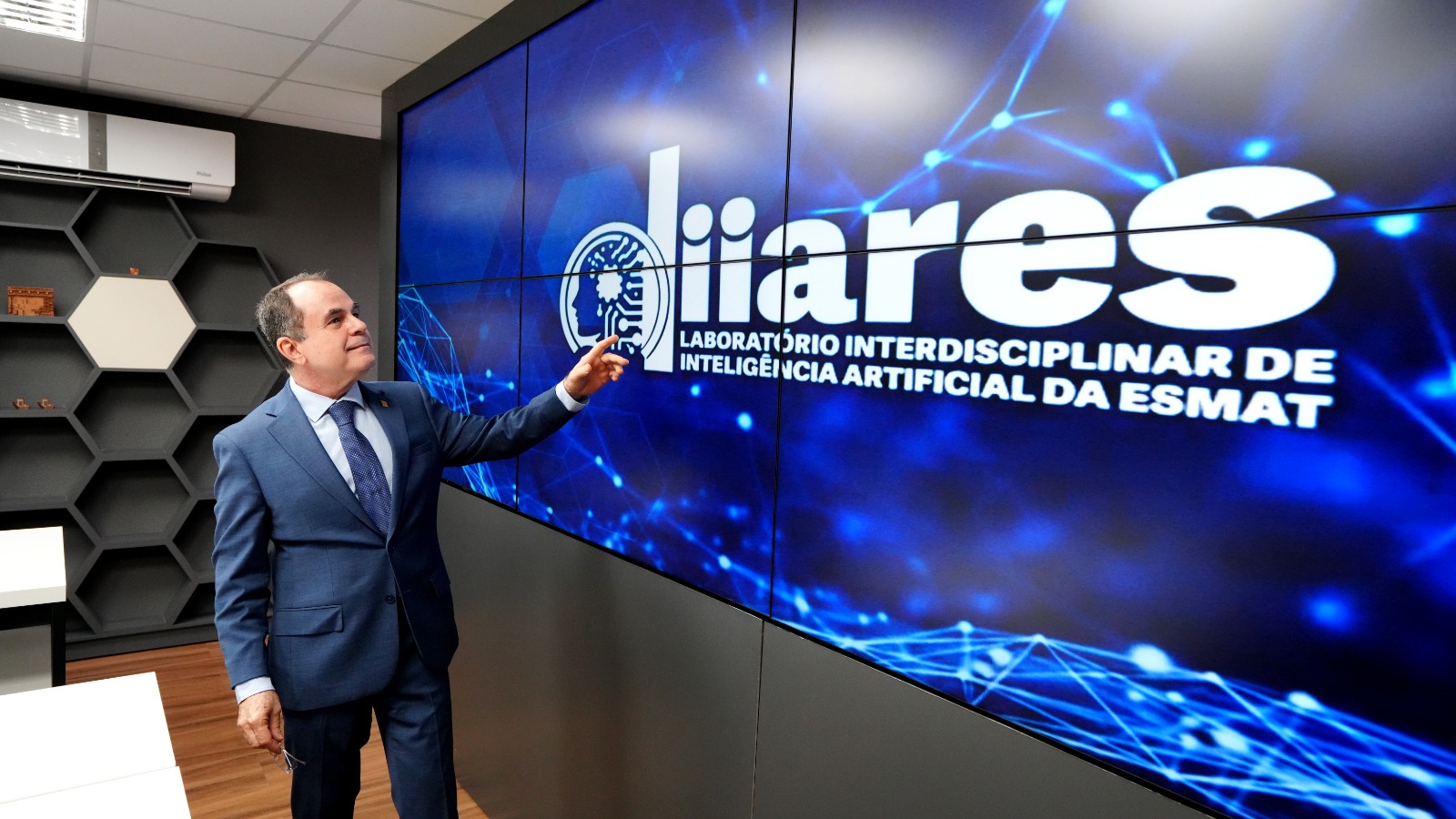 Homem de terno azul está em pé tocando uma grande tela digital com fundo azul e efeitos de conexões digitais.