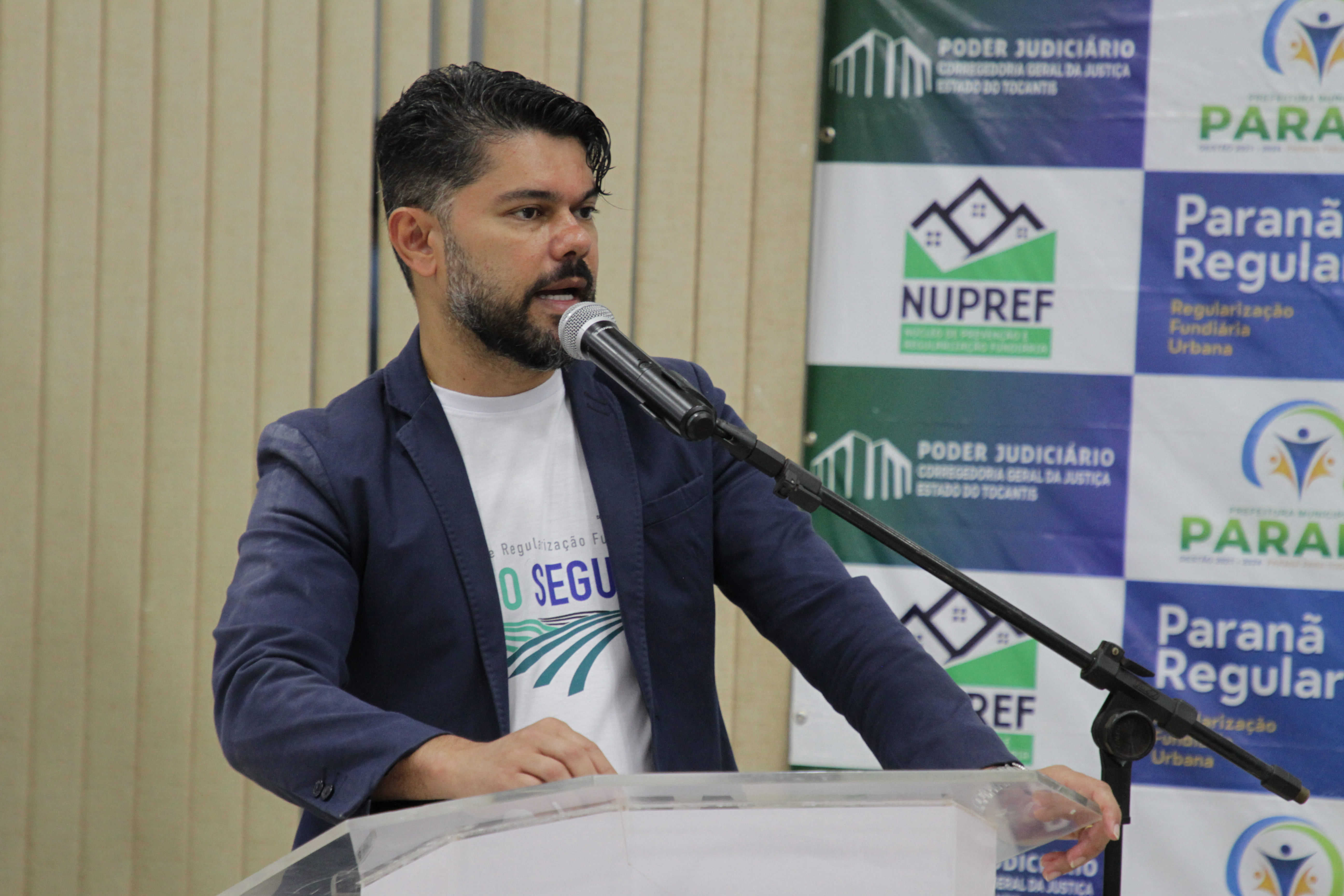 Homem de cabelo preto grisalho com blazer azul escuro e camisa branca com a palavra "Regularização Fundiária' estampada fala ao microfone