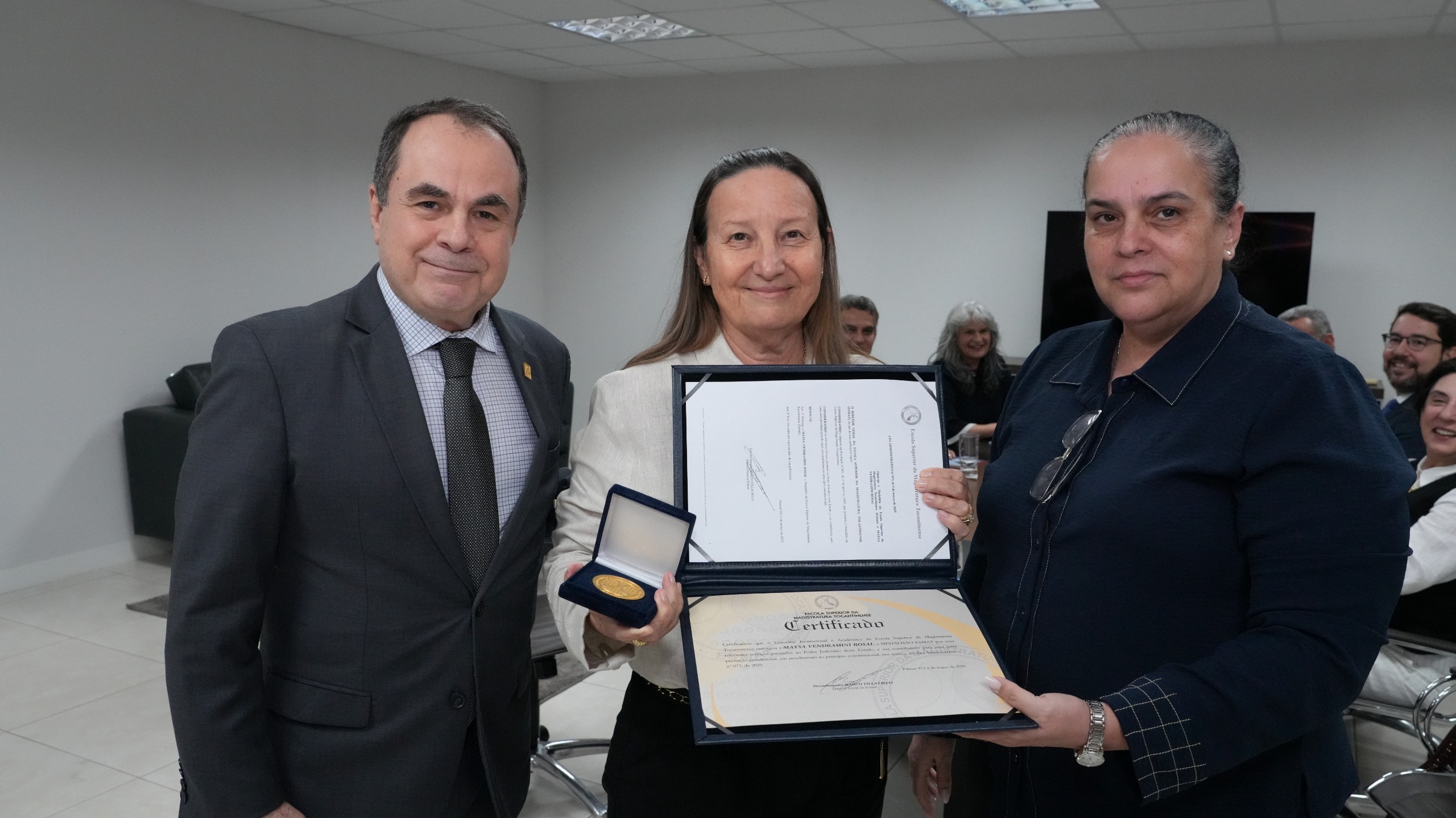 A imagem mostra três pessoas em uma sala de reunião bem iluminada, com paredes brancas e um teto com luminárias embutidas. Há cadeiras e algumas pessoas ao fundo, sentadas e sorrindo.    No centro, uma mulher de pele clara e cabelos longos e castanhos segura um certificado aberto e uma medalha dourada dentro de um estojo azul. Ela veste uma blusa clara.  À direita dela, há outra mulher de pele clara e cabelos presos. Ela veste uma camisa azul-escura e segura o certificado.  À esquerda da primeira mulher, um homem de pele clara, cabelos curtos e escuros, veste um terno cinza com uma gravata estampada. Ele tem uma expressão amigável e está olhando para a câmera.