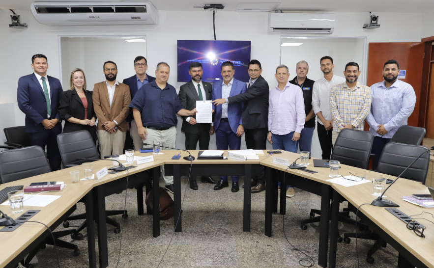magem institucional registra um grupo de cerca de 12 homens e uma mulher reunidos em uma sala de reuniões. Eles estão posicionados lado a lado, atrás de uma mesa em formato de “U”, equipada com microfones, copos de água e materiais de trabalho.  Ao centro, dois participantes seguram um documento
