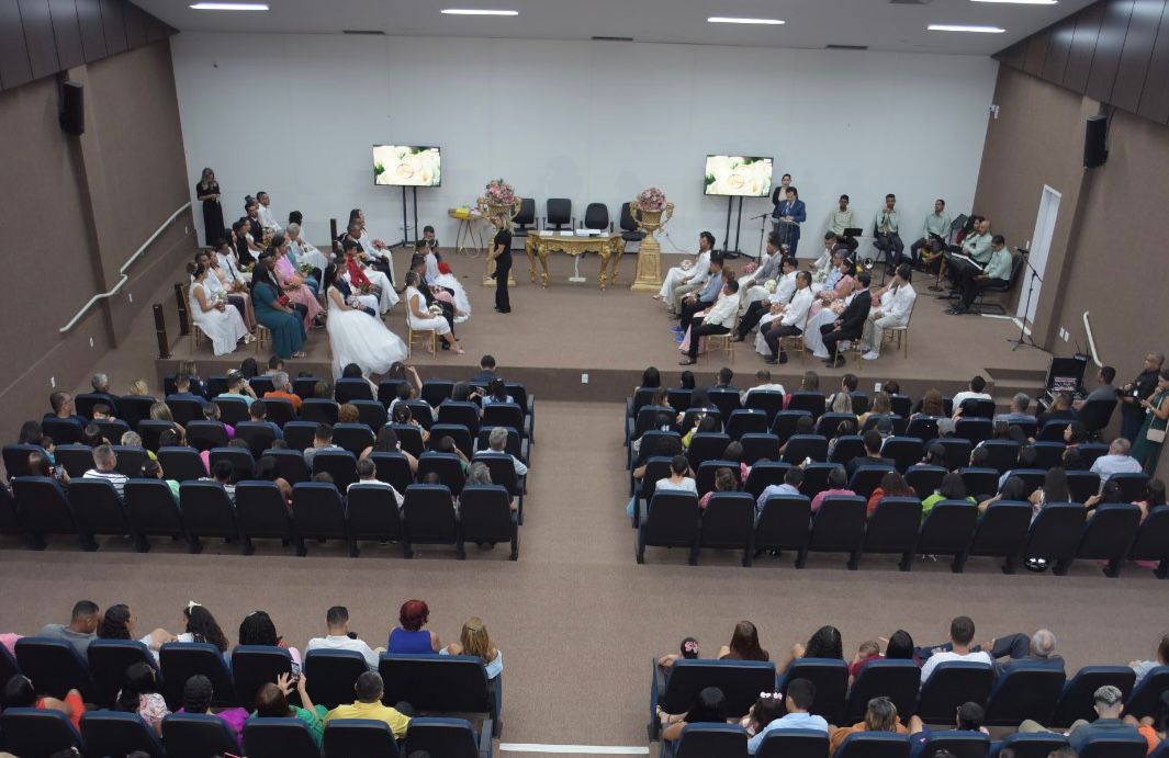 foto aberta retratando uma cerimonia de casamento coletivo; ao fundo os noivos e os covidados sentados no auditório
