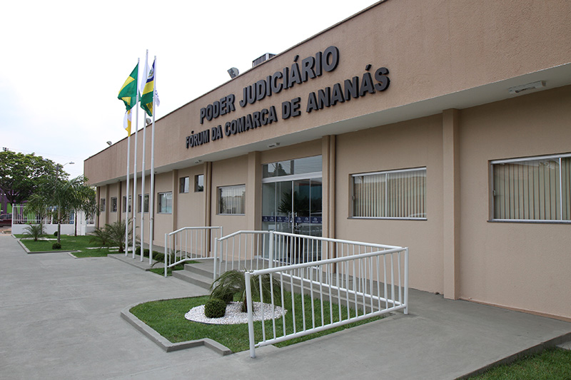 A imagem mostra a fachada do prédio do Fórum de Ananás, em tom bege claro, com janelas e portas bem distribuídas, além de rampas de acessibilidade com corrimãos metálicos. O entorno é bem cuidado, com gramado aparado e pequenas plantas que embelezam a entrada.