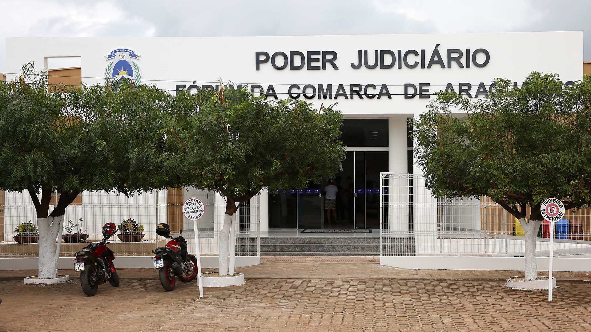 Fachada frontal na cor branca e o nome da comarca de Araguatins, com a visão do portão aberto, árvores na entrada, as colunas de sustentação e escada de acesso