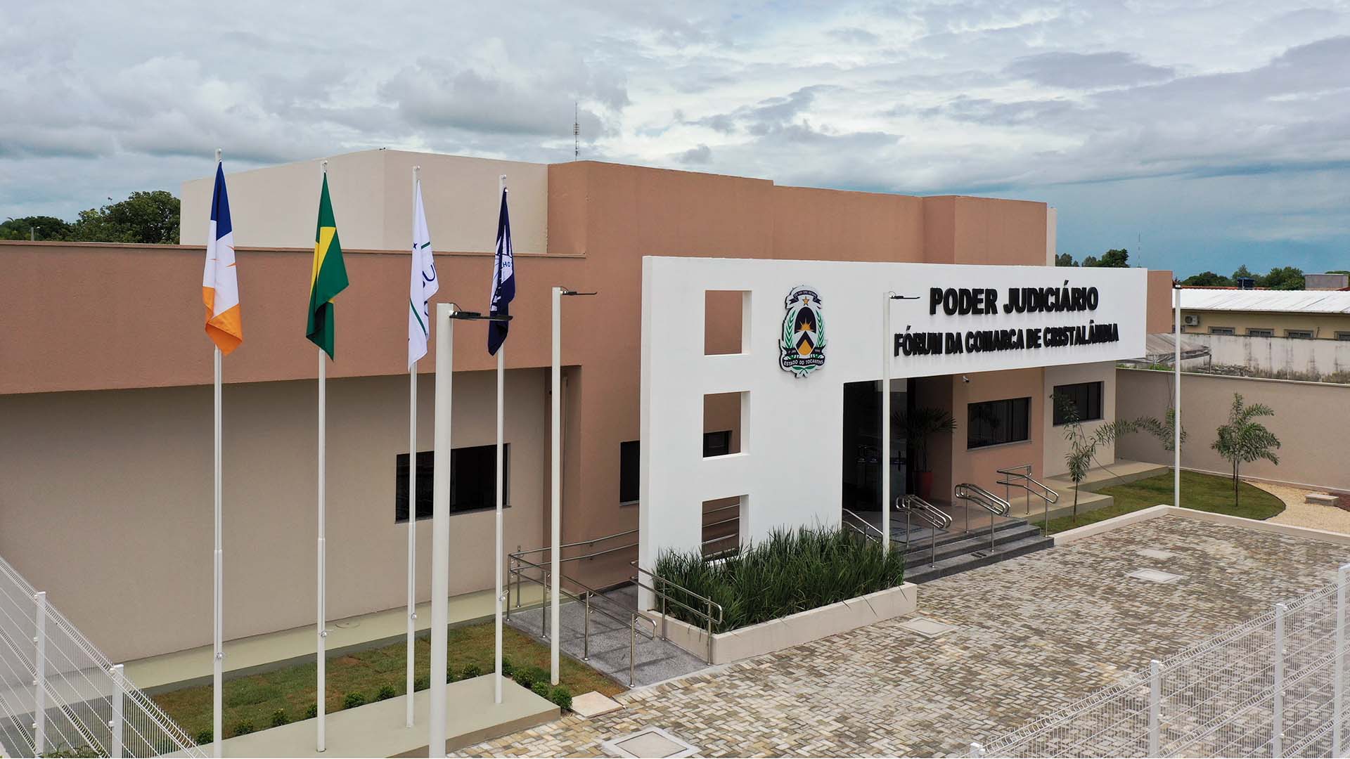 Fachada do Fórum de Cristalândia vista da perspectiva frontal e lateral, com parte do estacionamento, jardim frontal, mastros e bandeiras, o pórtico branco com letreiros na cor preta
