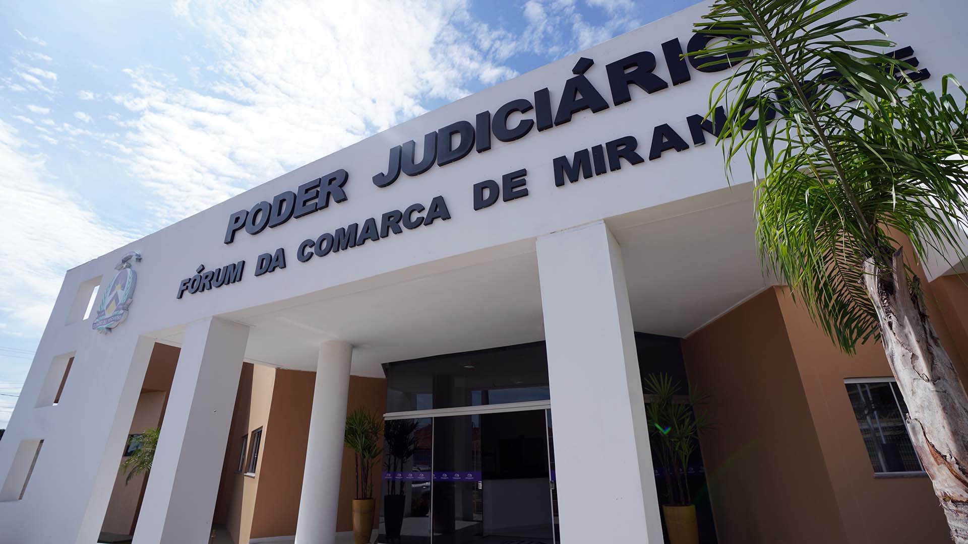 Fachada do prédio do Fórum de Miranorte, da perspectiva frontal, vista de baixo, com vista das colunas e fachada na cor branca, com letreiro do nome do fórum na cor preta, e paredes marrons
