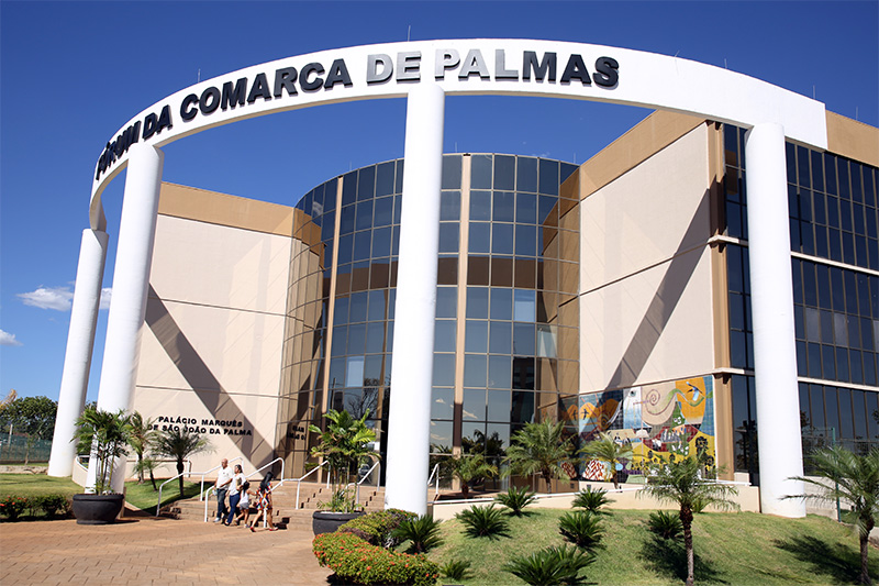 fachada do prédio da Comarca de Palmas; paredes nas cores bege e marrom, vidraças e um arco na parte frontal onde está o nome: Fórum da Comarca de Palmas; na frente do prédio um jardim 