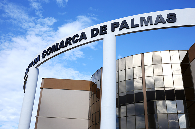 Detalhe da fachada do Fórum de Palmas com parte da parede espelhada em meio a dois detalhes das colunas da entrada principal e letreiro em cor preta no pórtico