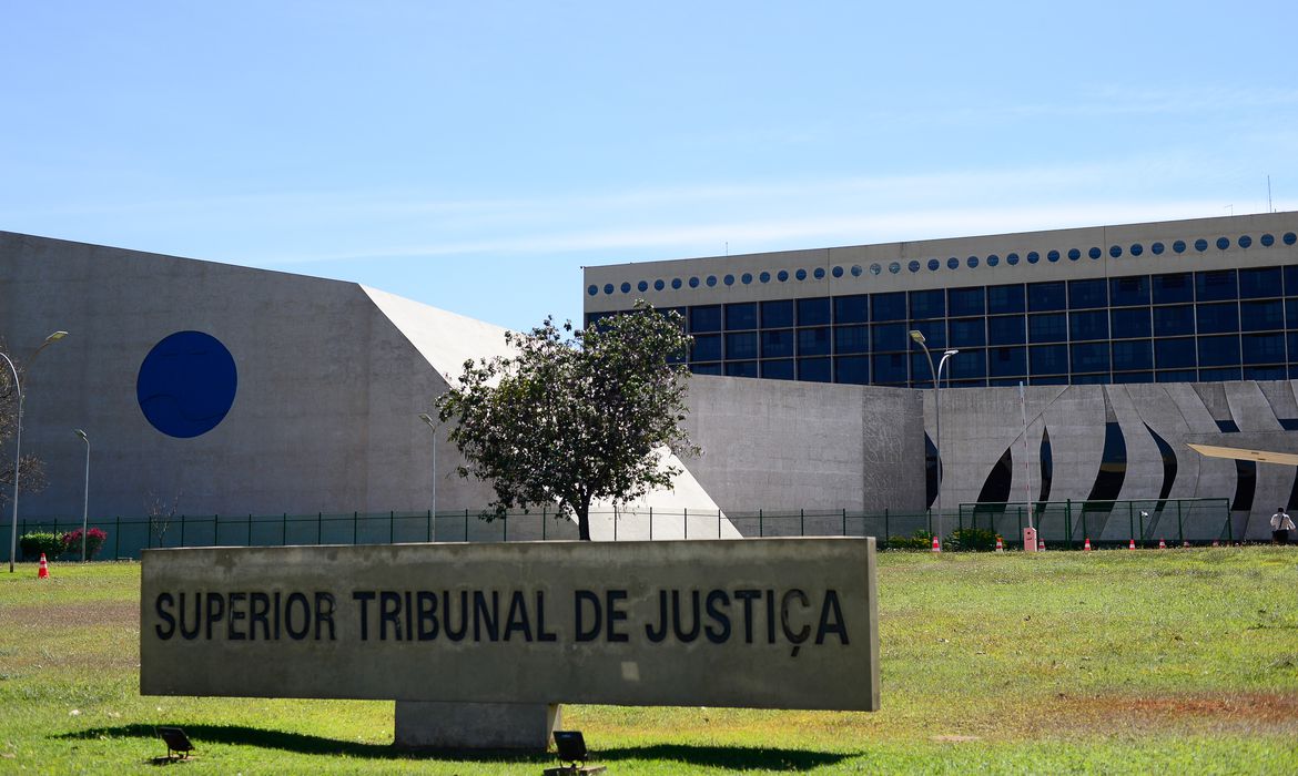 Imagem da fachada do prédio do Superior Tribunal de Justiça, em cor cinza, com vidraças escuras. à frente uma placa com o nome Superior Tribunal de Justiça