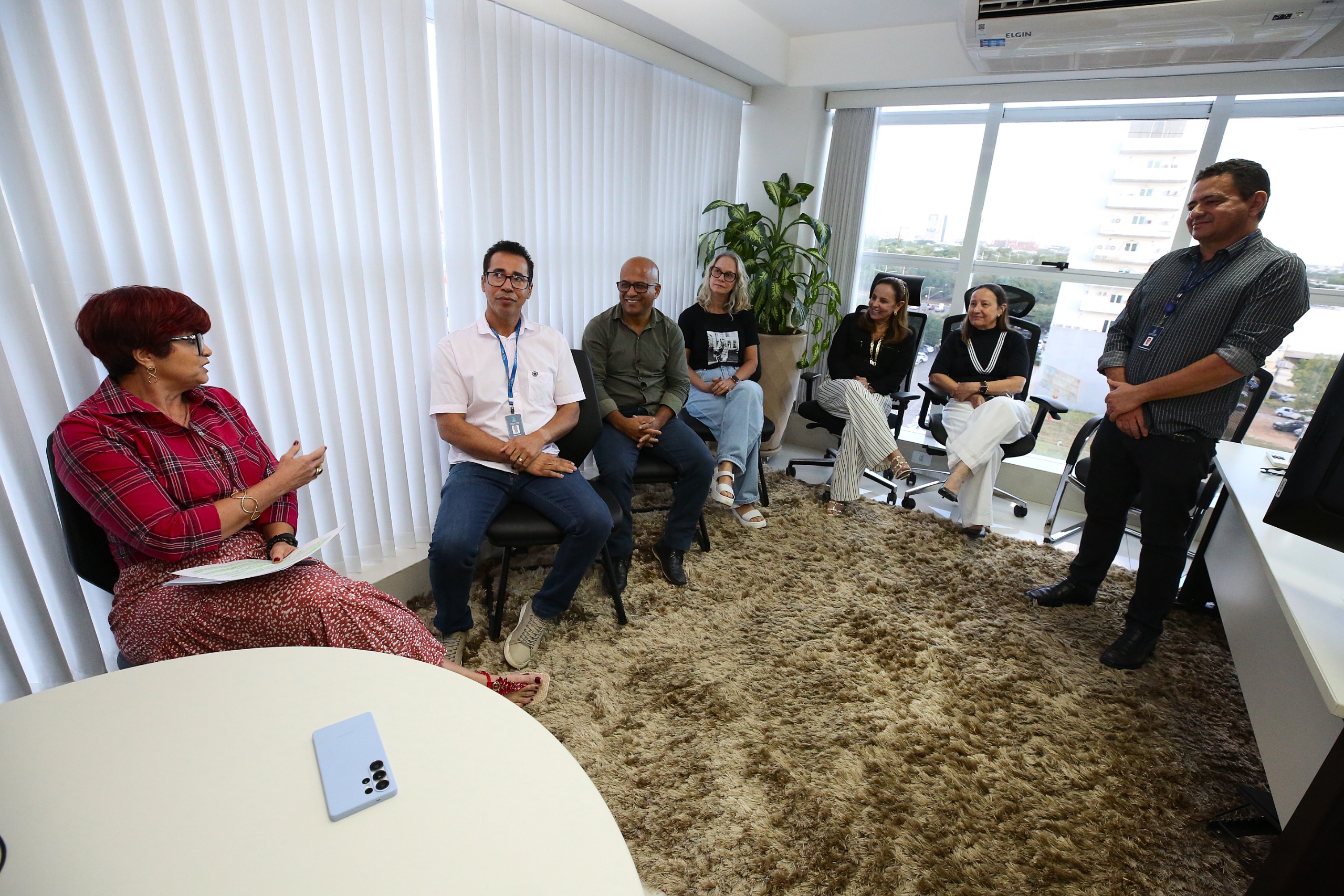 A imagem mostra um grupo de pessoas em uma sala de escritório, sentadas em cadeiras ao redor de uma mesa redonda. Há sete pessoas, sendo seis mulheres e um homem, todas vestindo roupas casuais de trabalho, como camisas e blazers. Elas parecem estar participando de uma reunião, com expressões atentas e concentradas.  A sala é bem iluminada, com uma grande janela ao fundo que oferece uma vista da cidade. O ambiente é moderno e acolhedor, com um carpete claro no chão. Na mesa, há um celular pequeno, mas não há muitos outros objetos visíveis.