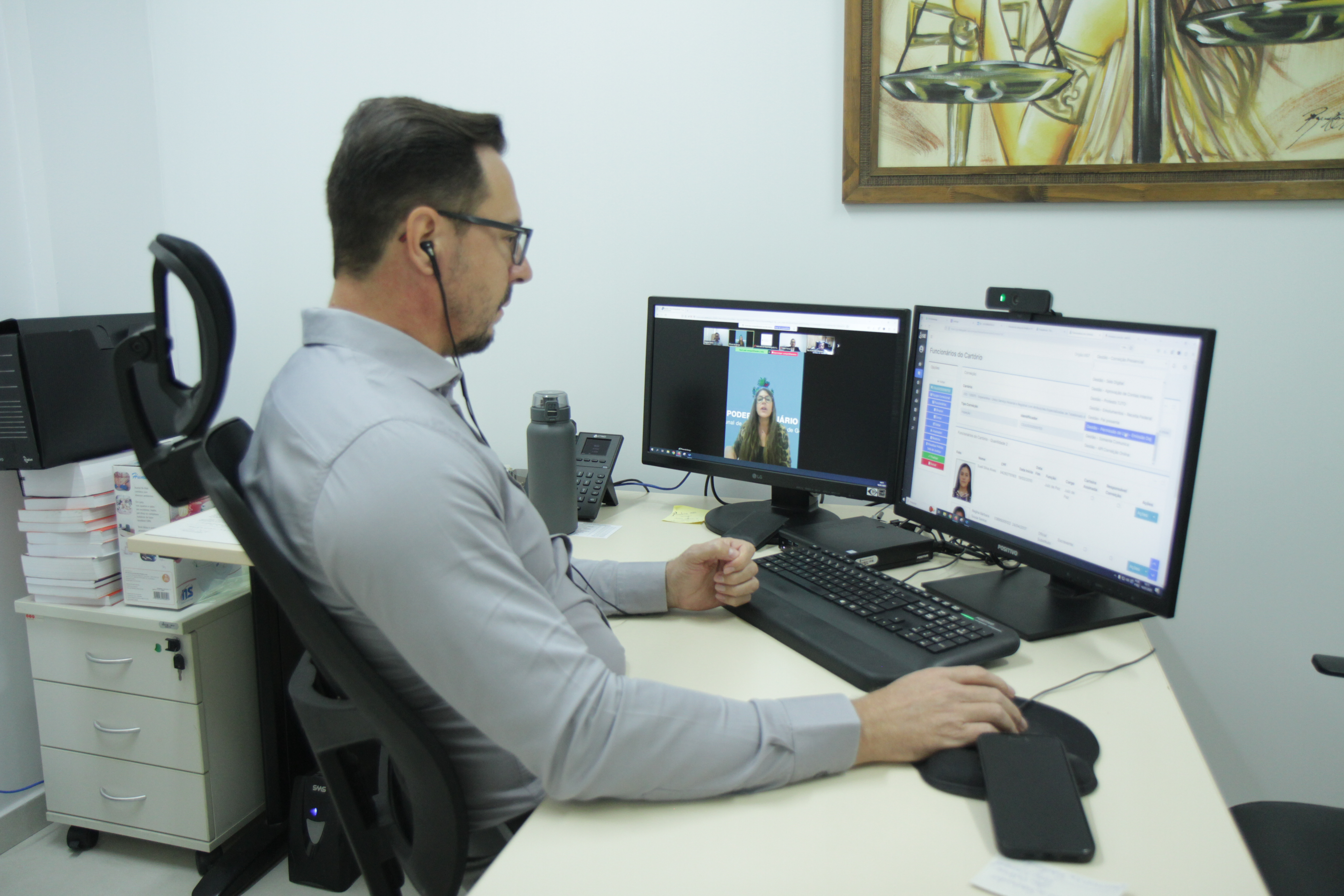 Homem de camisa social cinza e óculos sentado em frente a uma mesa que há dois monitores.