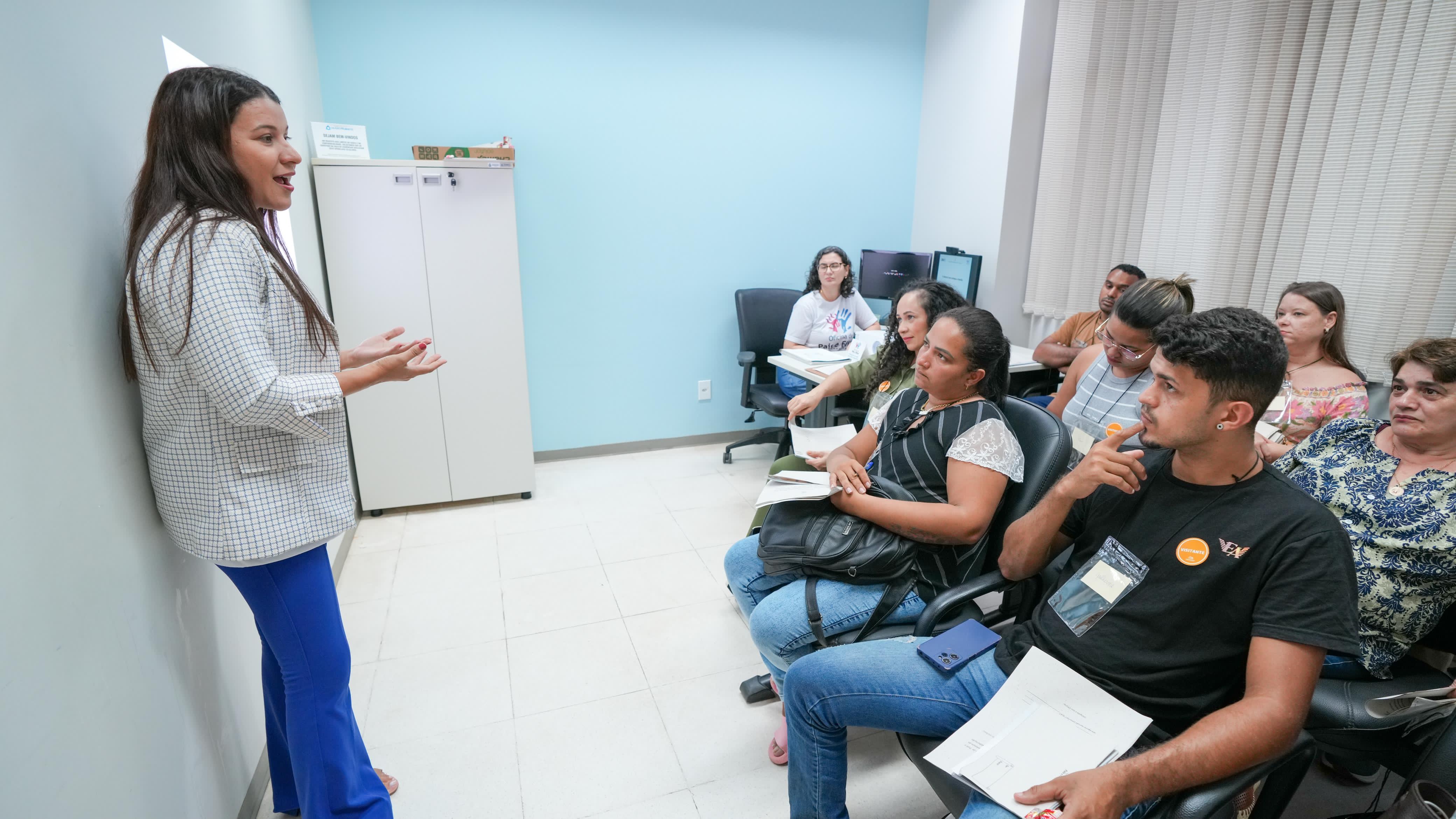 Sala na cor azul claro, mulher em pé do lado esquerdo, com um blazer xadrez e uma calça azul, o cabelo dela é preto e estar solto, ela gesticula com as mão como se estivesse conversando, ao fundo tem um armário branco, de  frente para mulher do lado direito tem um grupo de pessoas, homens e mulheres que estão olhando pra ele. 
