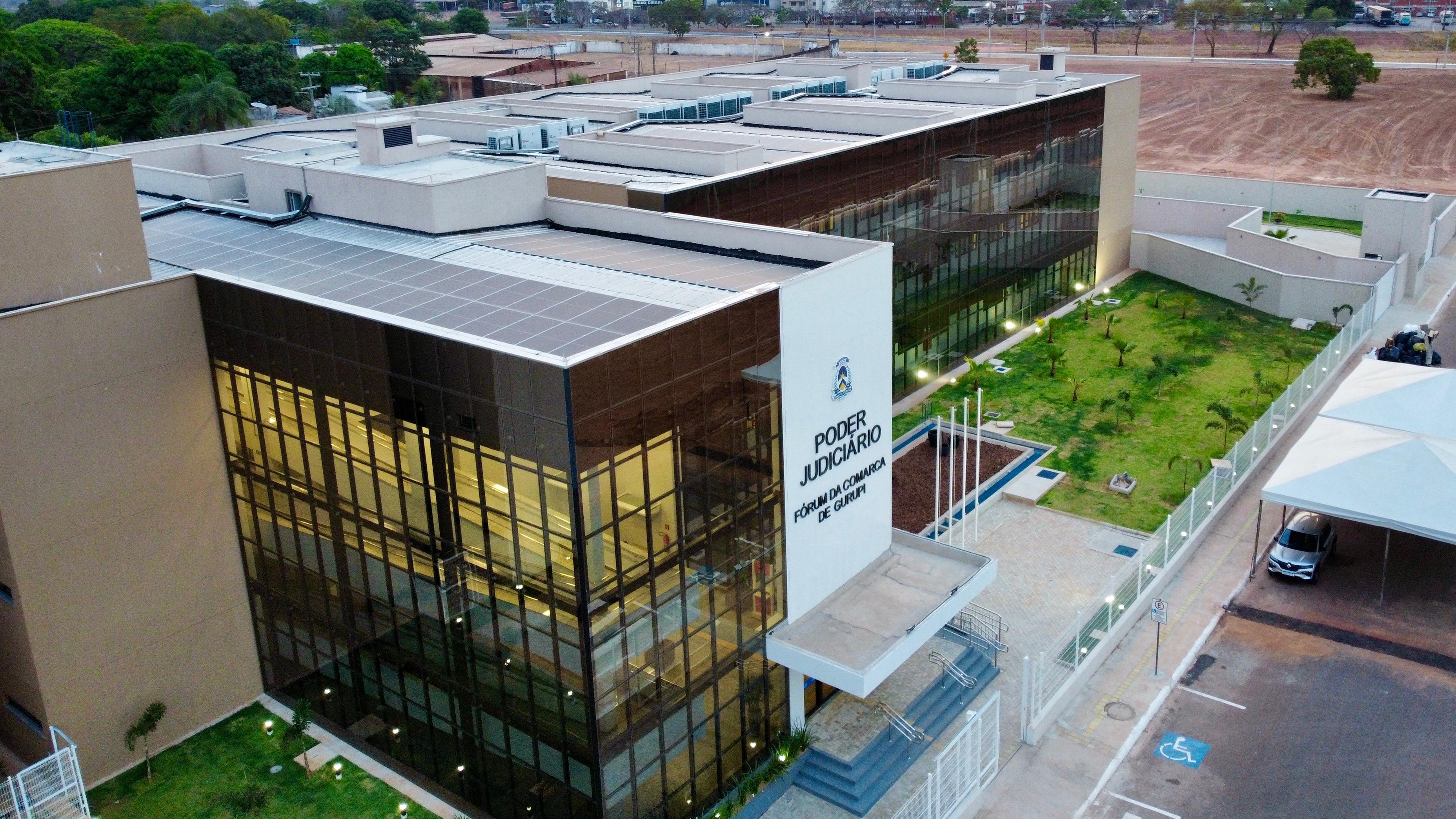 A foto mostra o prédio do Poder Judiciário do Tocantins, com paredes claras e vidros espelhados. Na frente tem uma placa com o nome, jardim ao lado e estacionamento na entrada.