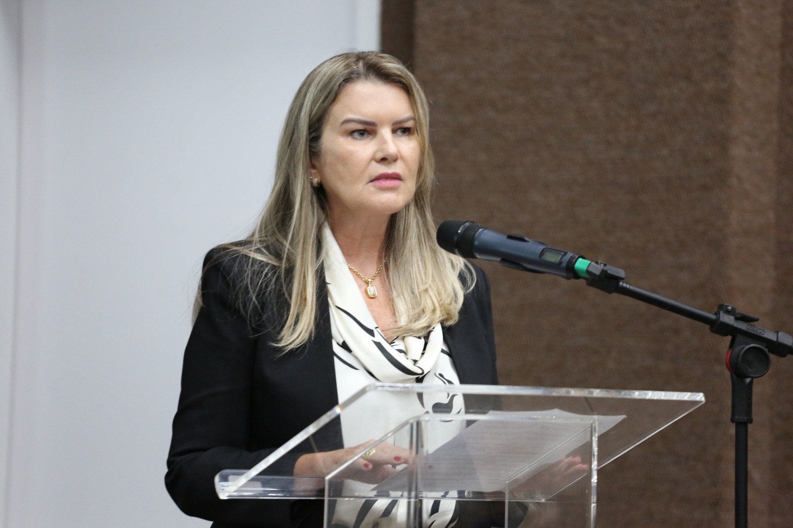 Mulher branca, loira com blazer preto e blusa branca com estampa fala ao microfone