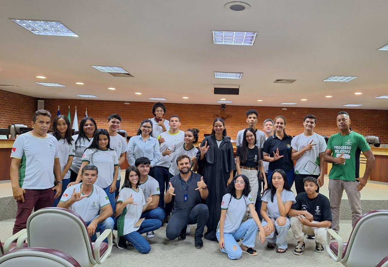 adolescentes posam para foto em auditório, ao centro uma aluna usa uma toga preta, entre eles estão a professora com blusa social branca, a intérprete de blusa preta, dois homens de apoio técnico, um com blusa verde e outro com blusa banca e ao centro um homem de blusa cinza, calça jens