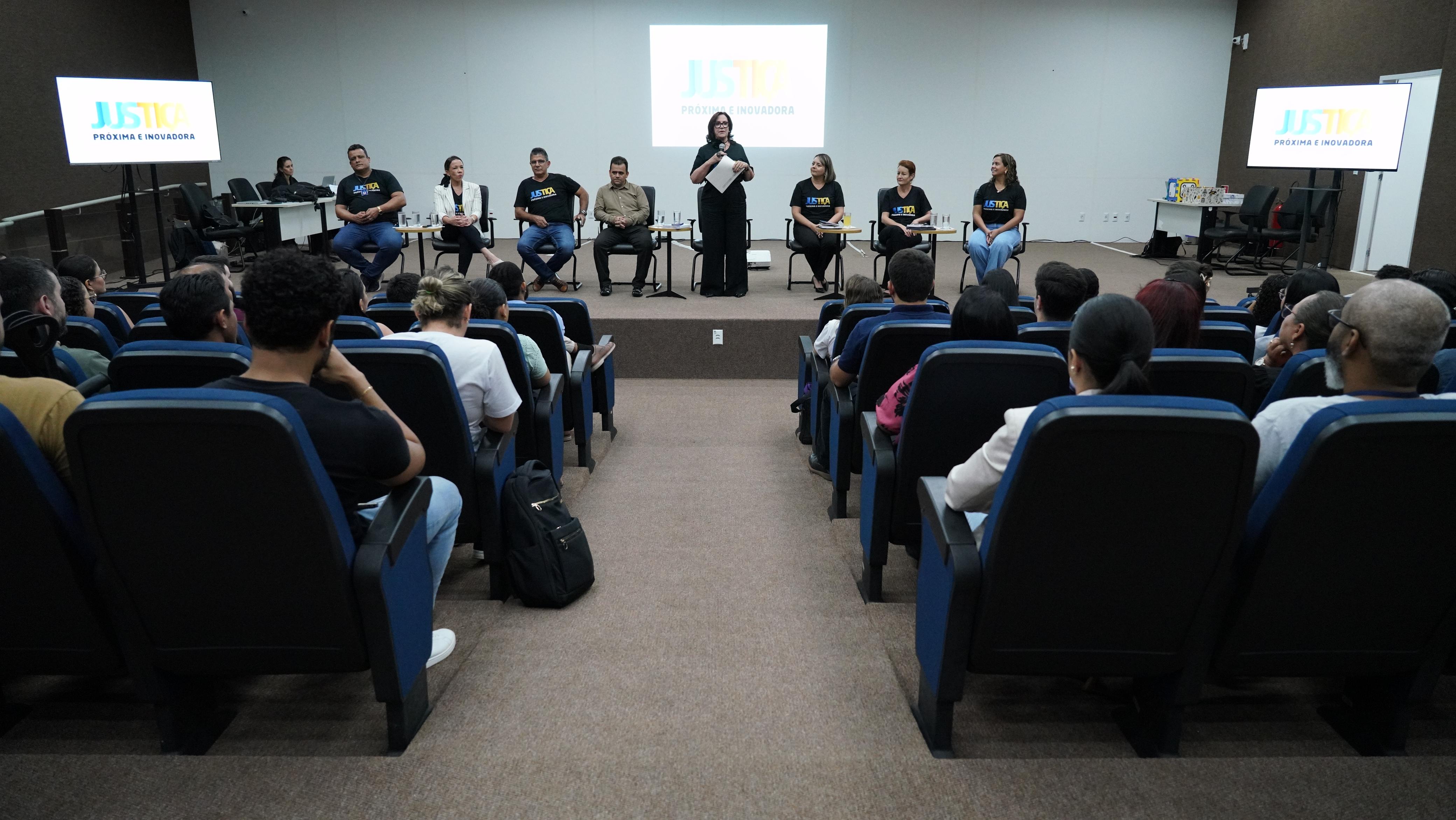 Grupo de pessoas sentadas em cadeiras azuis escutando atentamente a presidente do TJTO Etelvina que está posicionada no centro.