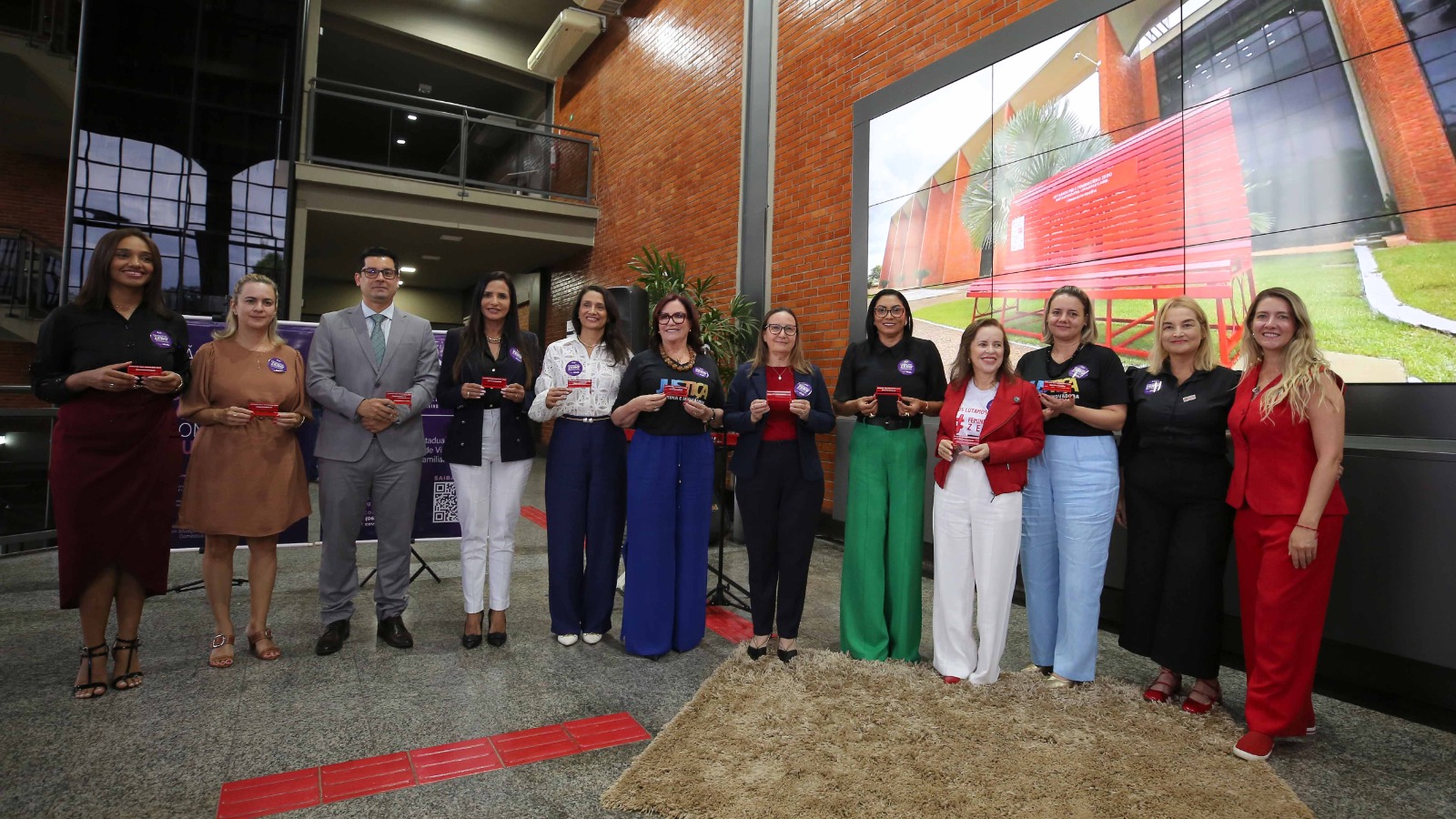 Autoridades, 11 mulheres e um homem, representantes de instituições que atuam na causa, posam para foto segurando uma miniatura do banco vermelho