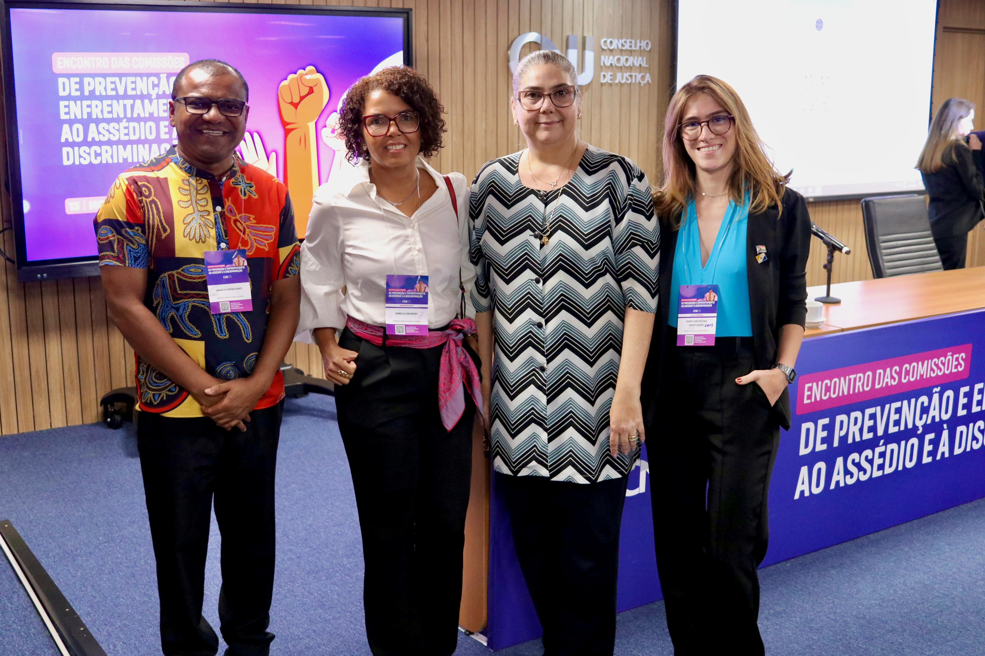 A imagem mostra quatro pessoas em pé, sorrindo, dentro de uma sala com decoração formal. Ao fundo, há uma tela com o título "Encontro das Comissões de Prevenção e Enfrentamento ao Assédio e à Discriminação" e o logotipo do Conselho Nacional de Justiça (CNJ). As pessoas usam crachás pendurados no pescoço e estão bem arrumadas, transmitindo um clima profissional e acolhedor.
