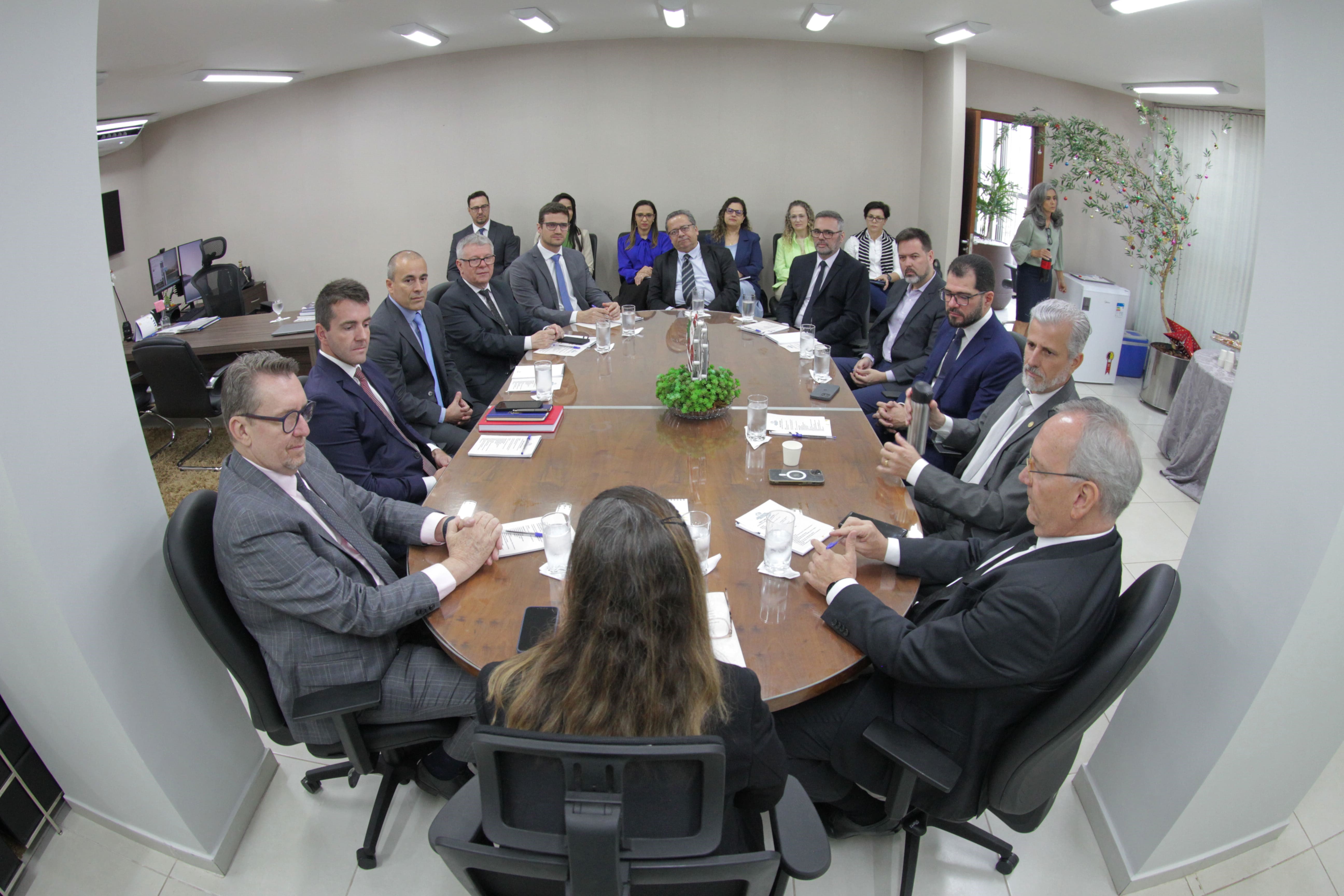 Grupo de pessoas vestidas formalmente e sentadas em torno de uma mesa grande e ao fundo, mais pessoas estão sentadas observando a reunião.