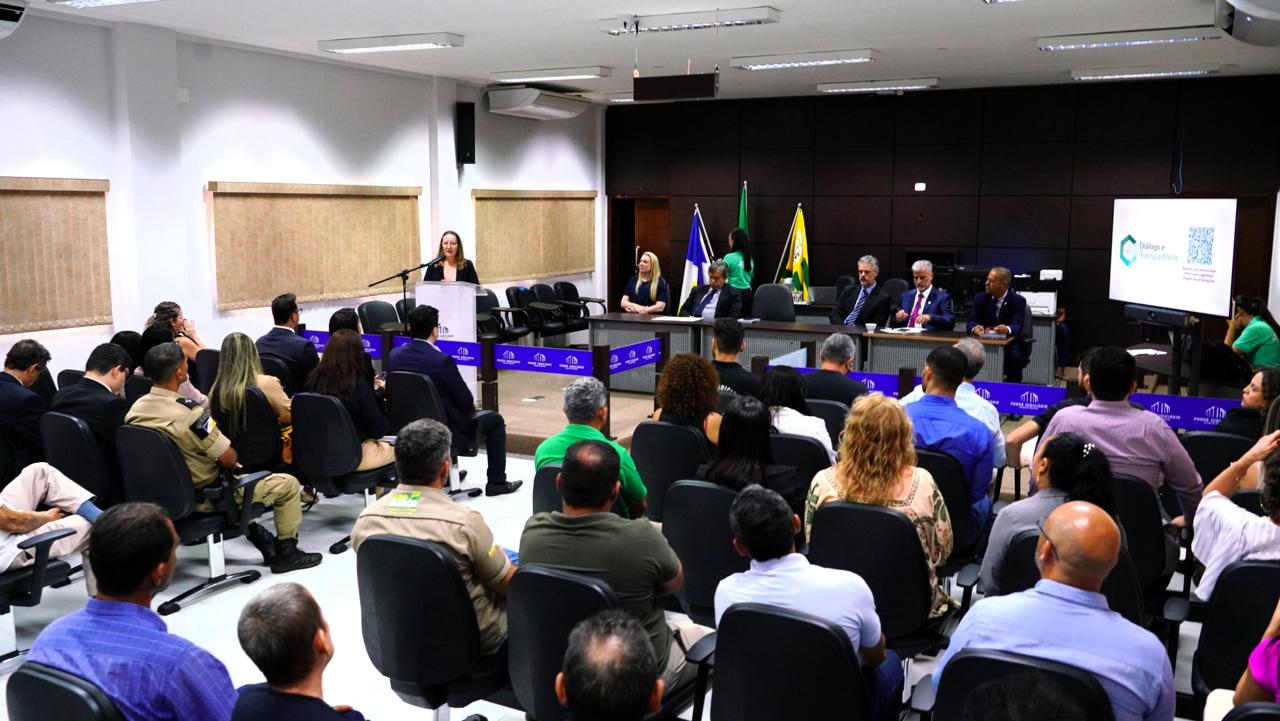 Grupo de pessoas reunidas em uma sala, sentadas em cadeiras pretas voltadas para frente que é composta por homens e mulheres.