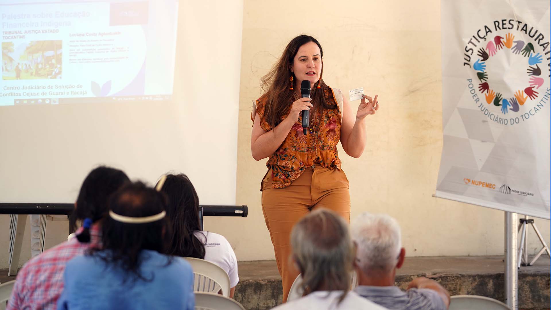 Juíza Luciana Aglantzakis em pé, usando calça e bluza caramelo com estampas coloridas, segurando um cartão e segurando um microfone. Ao lado do banner com a logo da Justiça Restaurativa