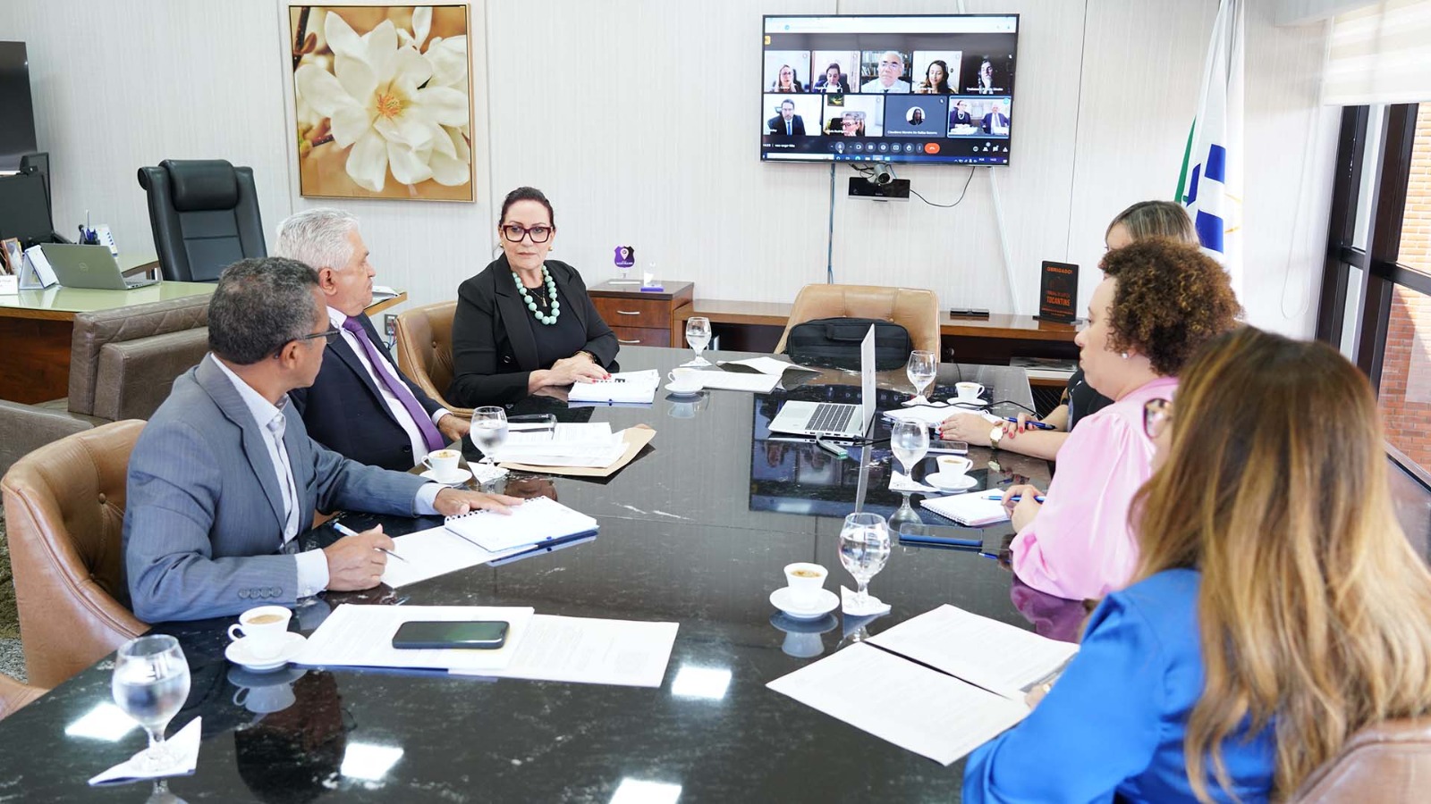 Presidente do TJ reunida em uma mesa com desembargador Lamounier, juíza Rosa Maria, diretora Ana Carina, chefe de gabinete Jeane; ao fundo um monitor na parede com demais participantes da comissão concurso e da FGV
