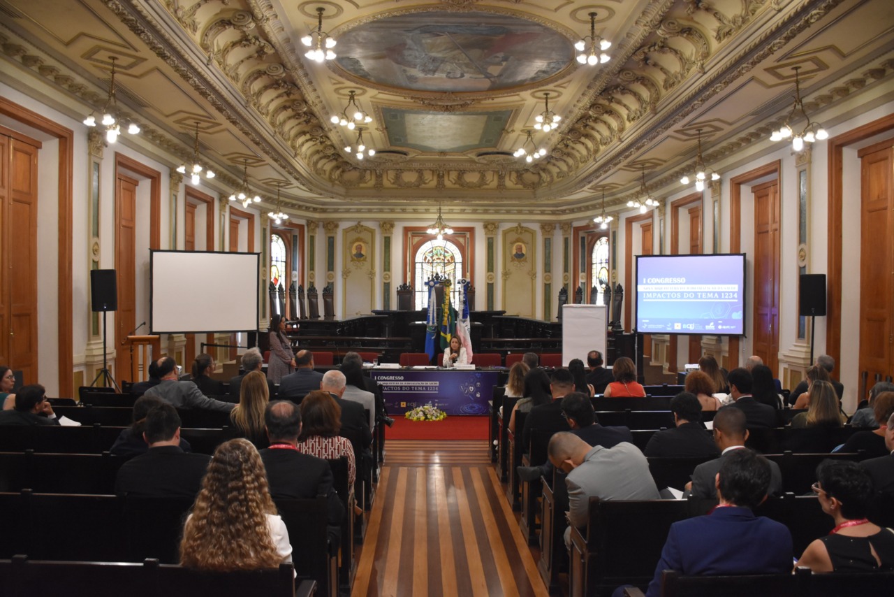 Foto aberta; na imagem um auditório com o público assistindo a uma palestra; ao fundo a palestrante sentada numa mesa de conferência, falando ao microfone