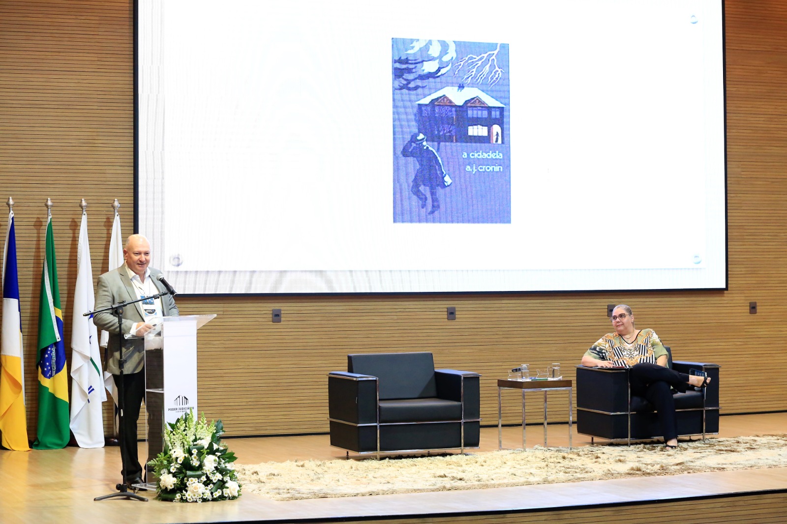 Em um palco, um palestrante, um senhor branco de terno bege, camisa branca e calça preta, fala ao microfone; à direita, sentada em uma das duas poltronas pretas que compõem o cenário, a juíza Flávia Bovo, uma senhora de blusa estampada e calça preta. Ao fundo, bandeiras do tocantins, Brasil e um telão