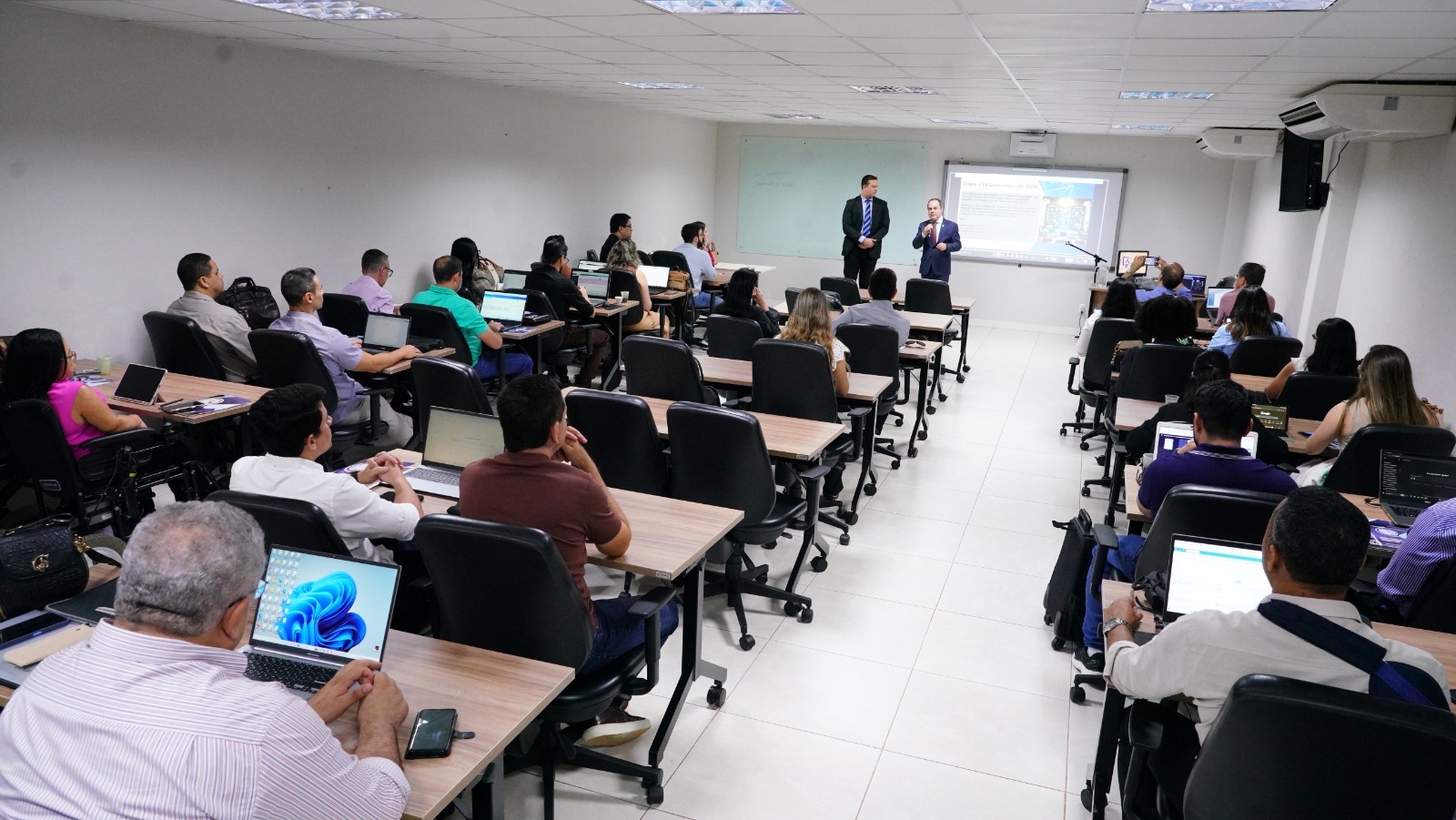 Foto de uma sala de aula com muitos alunos; eles estão sentados, posicionados de costa, assistindo o instrutor do curso e o diretor da Esmat falarem