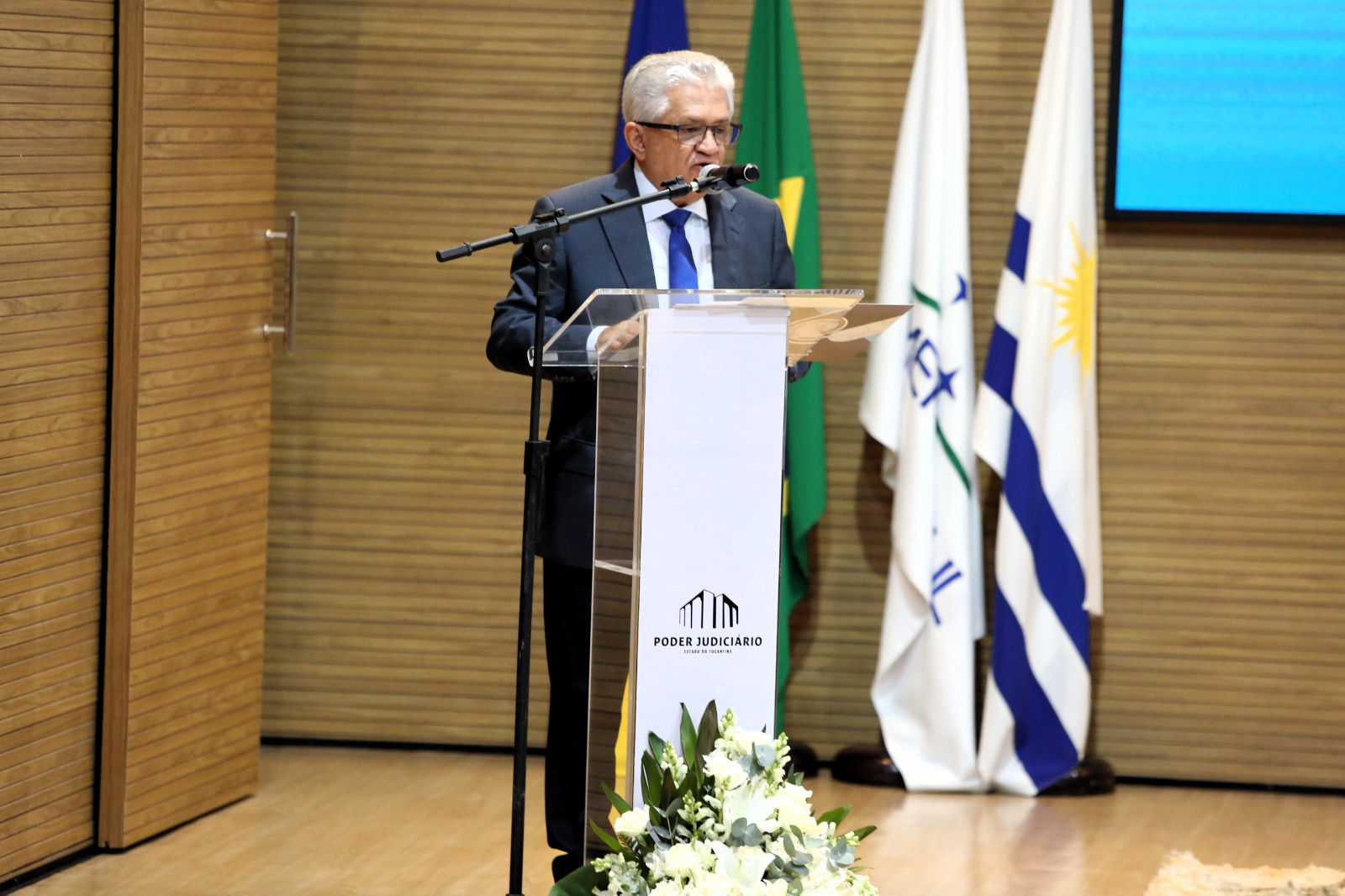 Homem grisalho com terno preto fala ao microfone emcima de palco em madeira