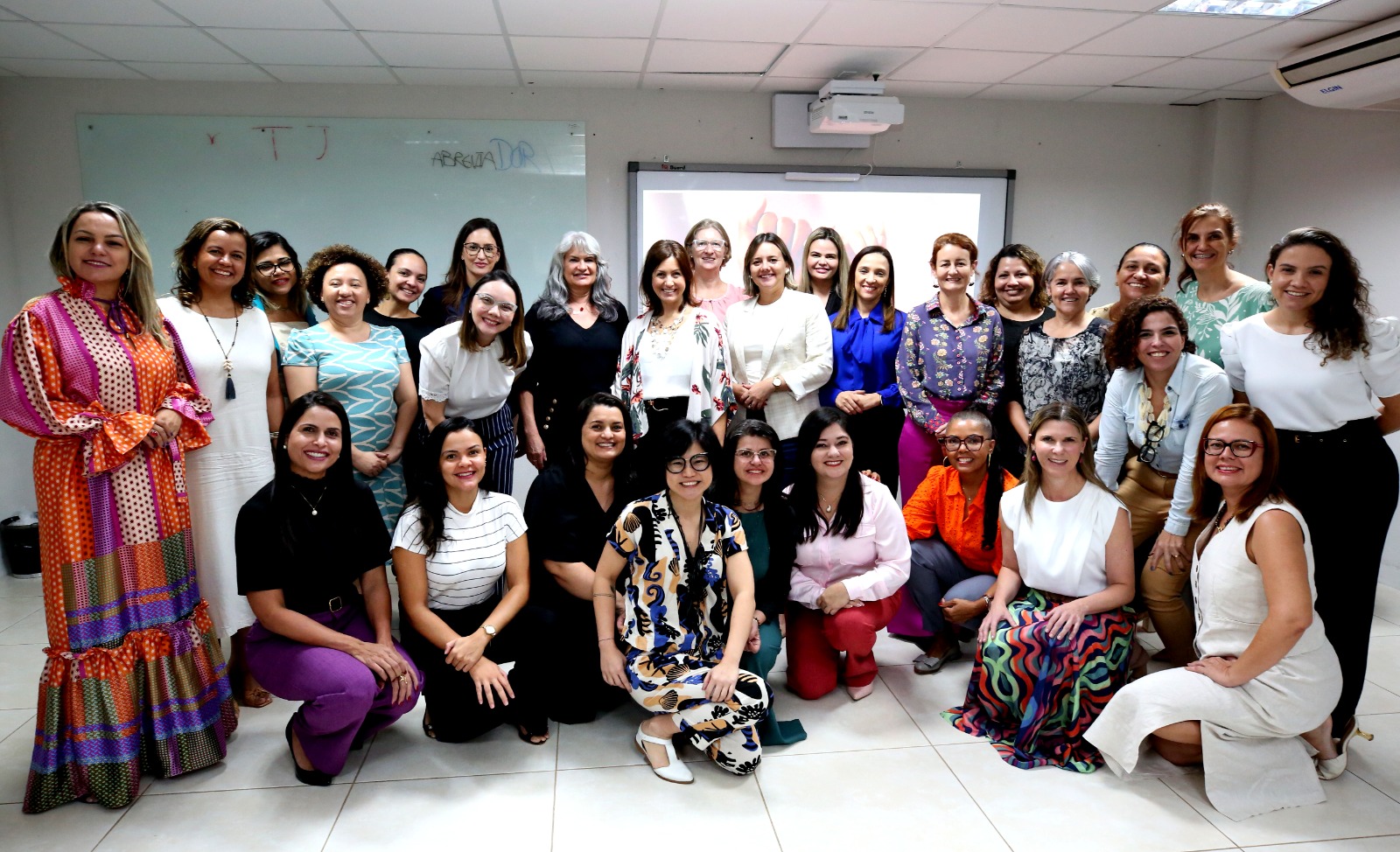 Grupo de pessoas posando para foto, algumas estão em pé, enquanto outras estão agachadas em um piso branco. Ao fundo há um quadro branco e uma tela de projetor que exibe informações.