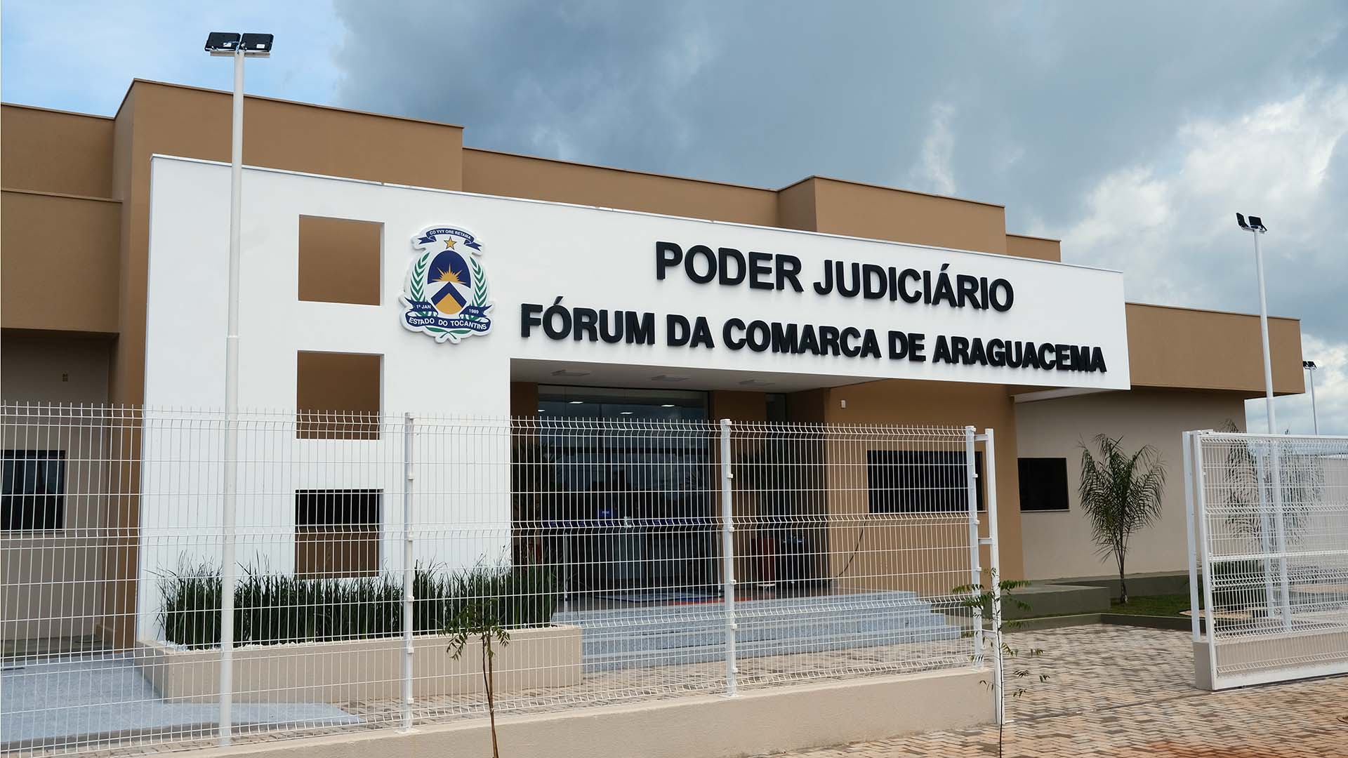 A imagem mostra a fachada de um prédio de justiça.  Na parte superior, está escrito em letras grandes "PODER JUDICIÁRIO" e abaixo "FÓRUM DA COMARCA DE ARAGUACEMA". Há um escudo com o brasão do Estado do Tocantins. O prédio é de cor bege e branco, cercado por uma cerca de metal branca.  Há postes de iluminação ao lado do prédio.