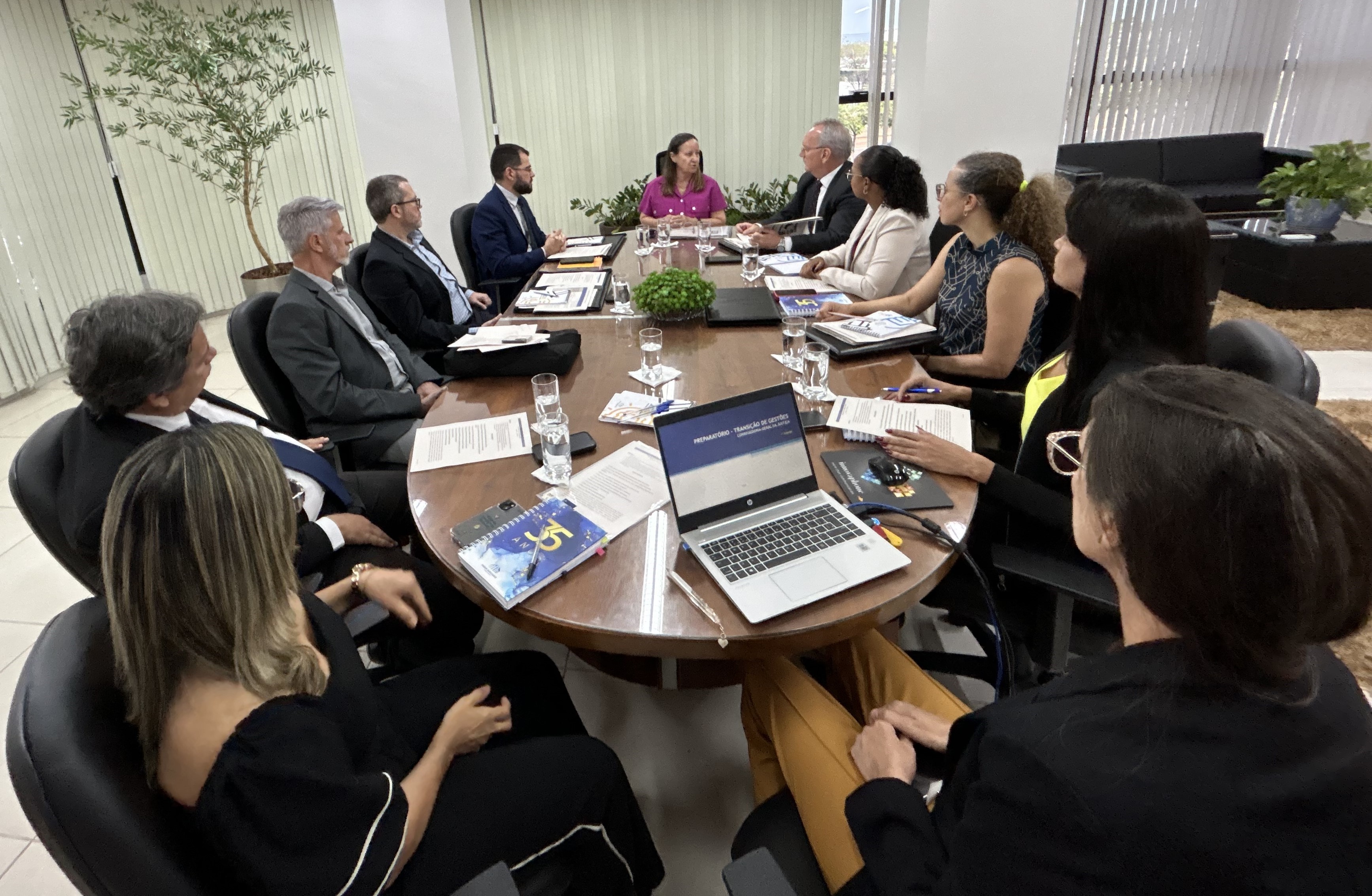Grupo de 11 pessoas reunidas, sentadas em volta de uma mesa, estão vestidos formalmente e atentos.