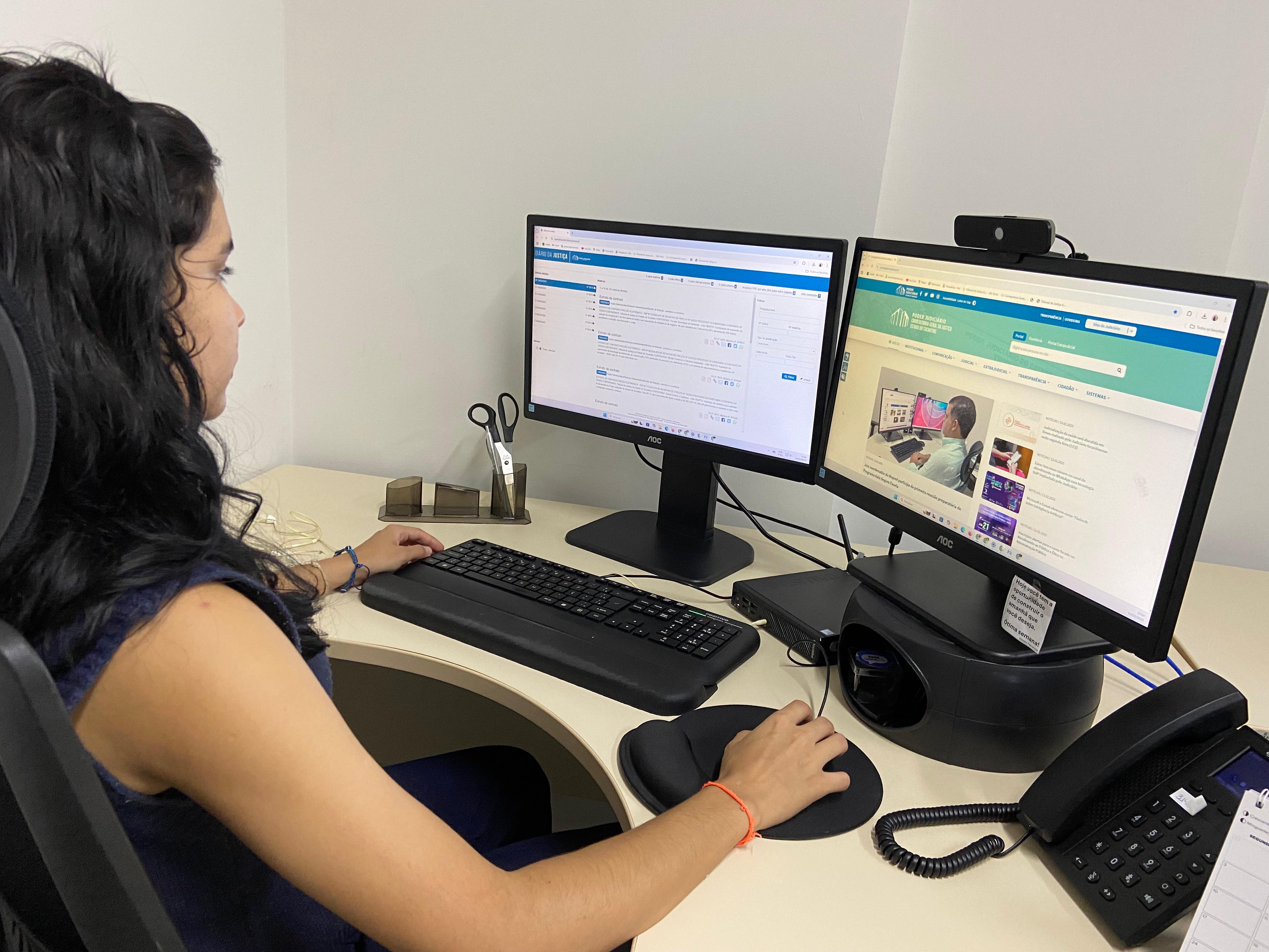 Mulher sentada próximo a mesa branca de computador, passando a ideia que estar olhando para a página do computador, ela segura no mouse, na sua frente tem duas telas