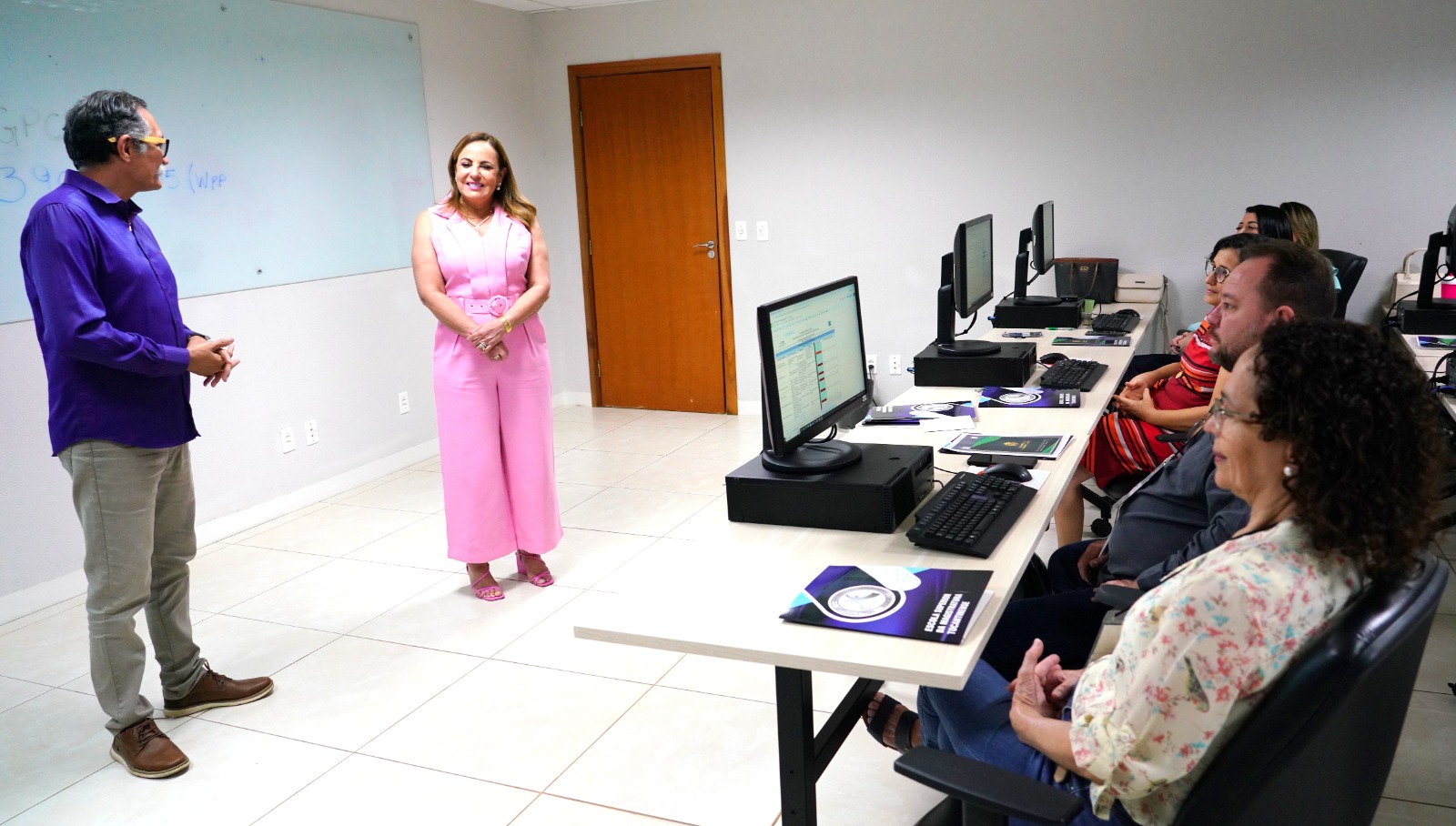 Desembargadora Ângela Prudente, em pé, vestindo um macacão rosa, ao lado do facilitador do Gestcom, em pé, à esquerda, com uma camisa roxa e claça bege, falando aos servidores, que estão sentados em frente a computadores