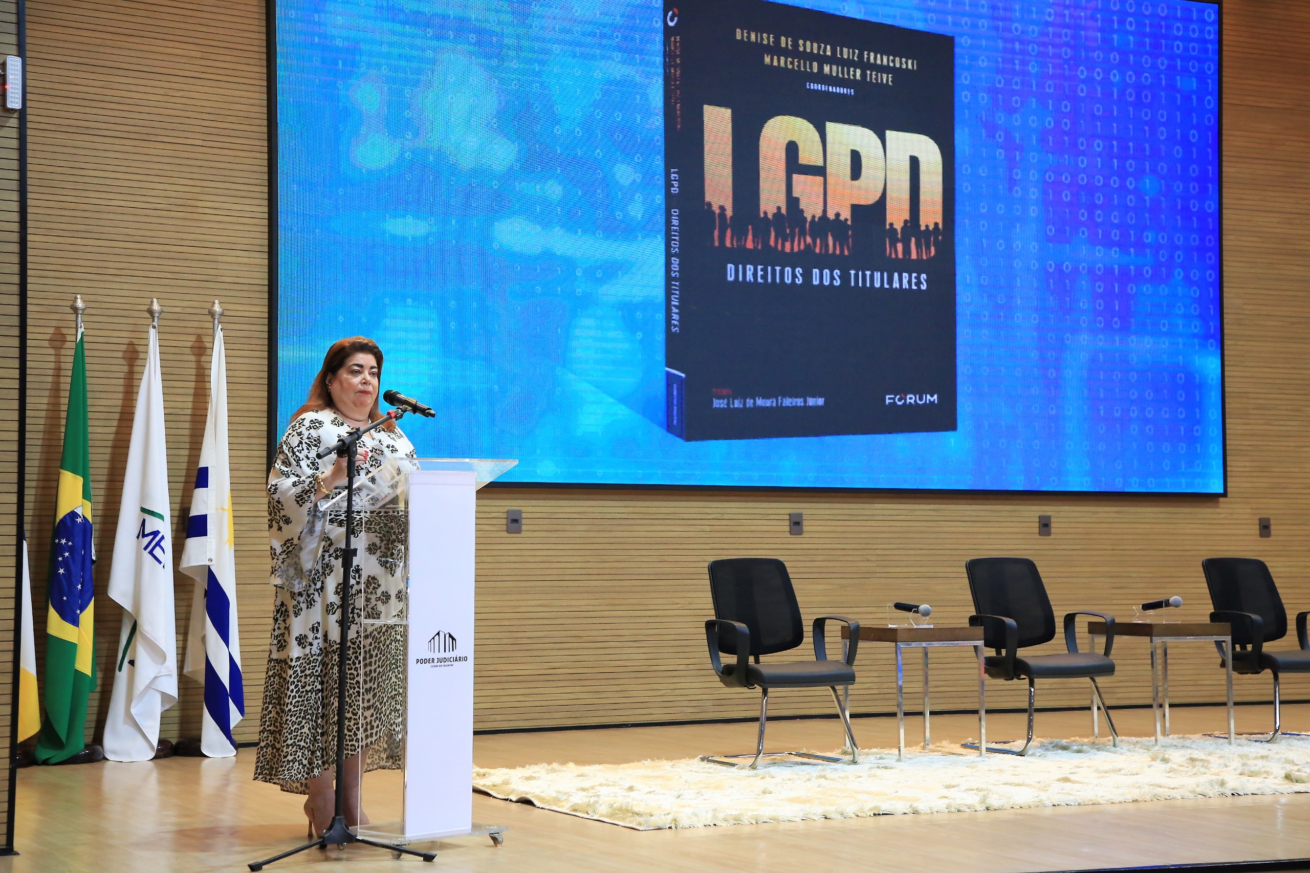 desembargadora denise, uma mulher de cabelos longos, usando vestido branco estampado em tons de marrom e preto, fala ao microfone em um pulpito à direita do palco do auditório
