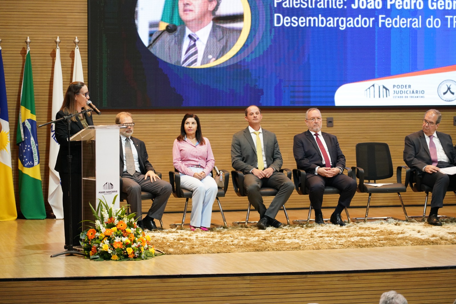 Imagem aberta da tribuna de honra com as autoridades presentes no evento; à esquerda, no púlpito, a presidente do TJTO, uma mulher banca, de cabelos castanhos, roupa escura e óculos, fala ao microfone ao centro, as autoridades sentadas e ao fundo um painel com a imagem dos palestrantes 