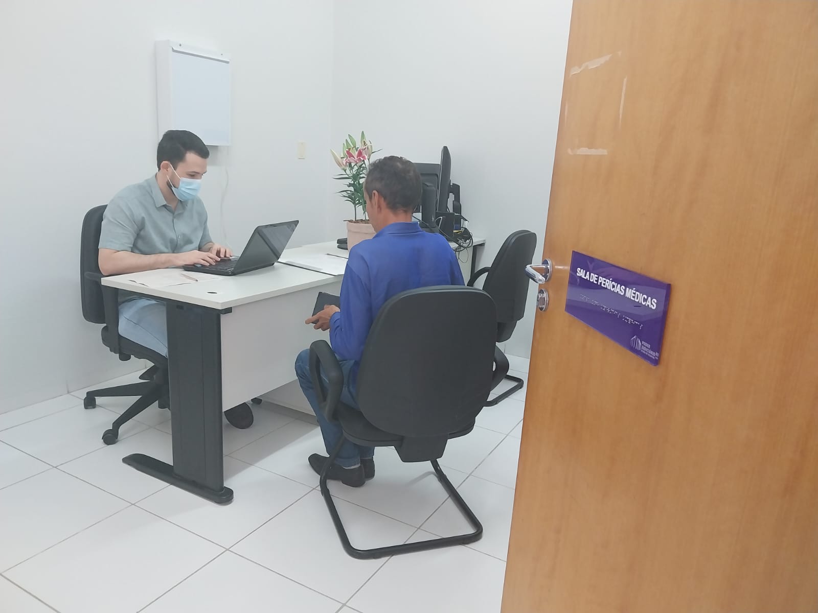 médico atendendo um paciente para perícia médica previdenciária; o médico está sentado, usando um notebook e o paciente, um homem, está sentado de costa; em primeiro plano uma porta aberta de madeira com uma placa azul escrita  Sala de Perícias Médicas em fonte branca