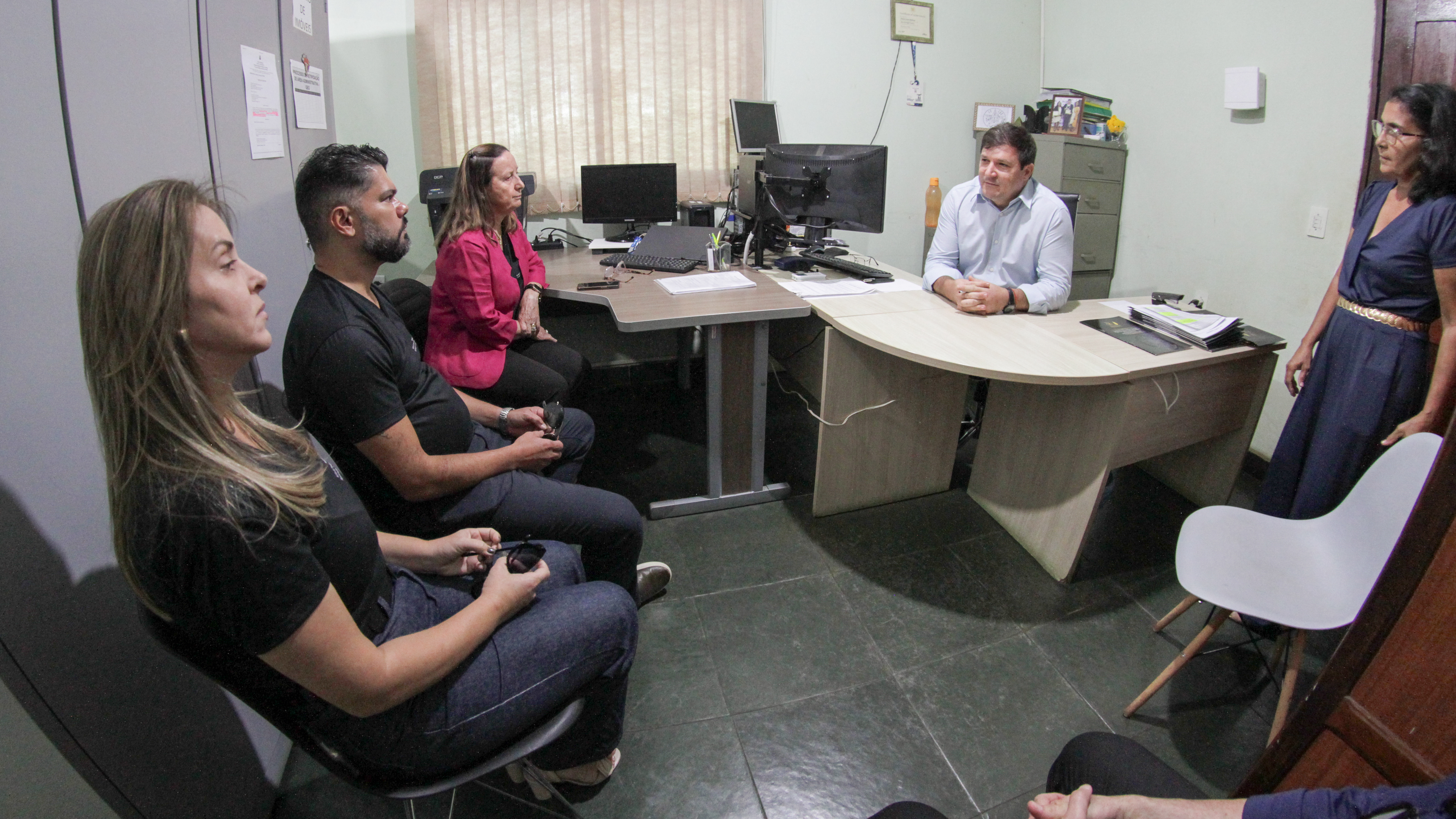 cinco pessoas em uma sala, três delas sentadas a frente de um homem e ao seu lado uma mulher que o observa