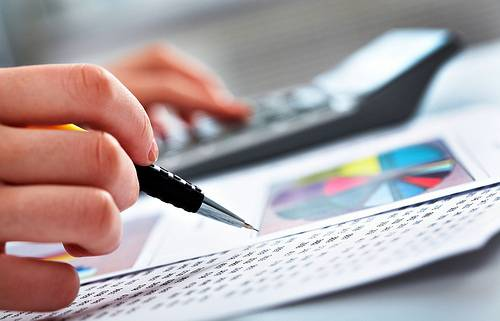 Imagem ilustrativa do trabalho de controladoria mostra uma mão segurando lapiseira que aponta para gráficos e tabelas impressos sobre a mesa